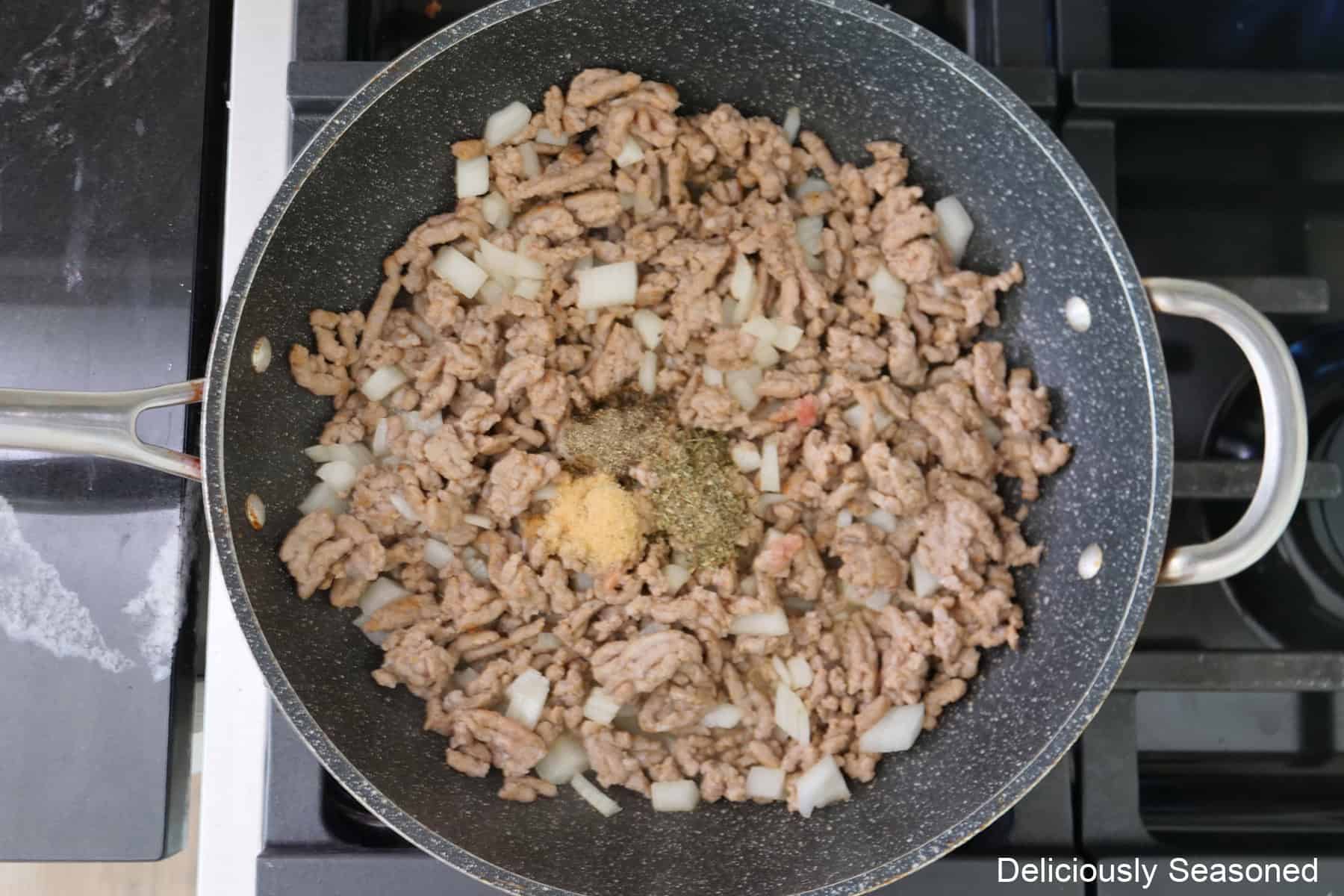 A frying pan on the stove with cooked sausage, onions, and seasonings in it.