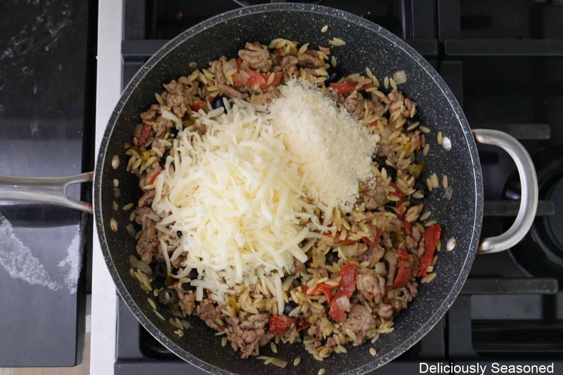 A frying pan on the stove with the sausage mixture and shredded mozzarella and Parmesan cheese added on top before being mixed into the mixture.