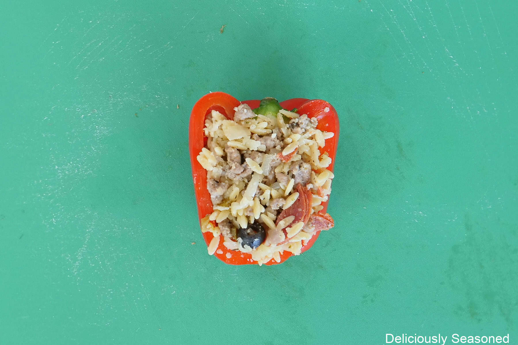 A green cutting board with a red bell pepper that has been cut in half and stuffed with pepperoni pizza orzo sausage mixture.