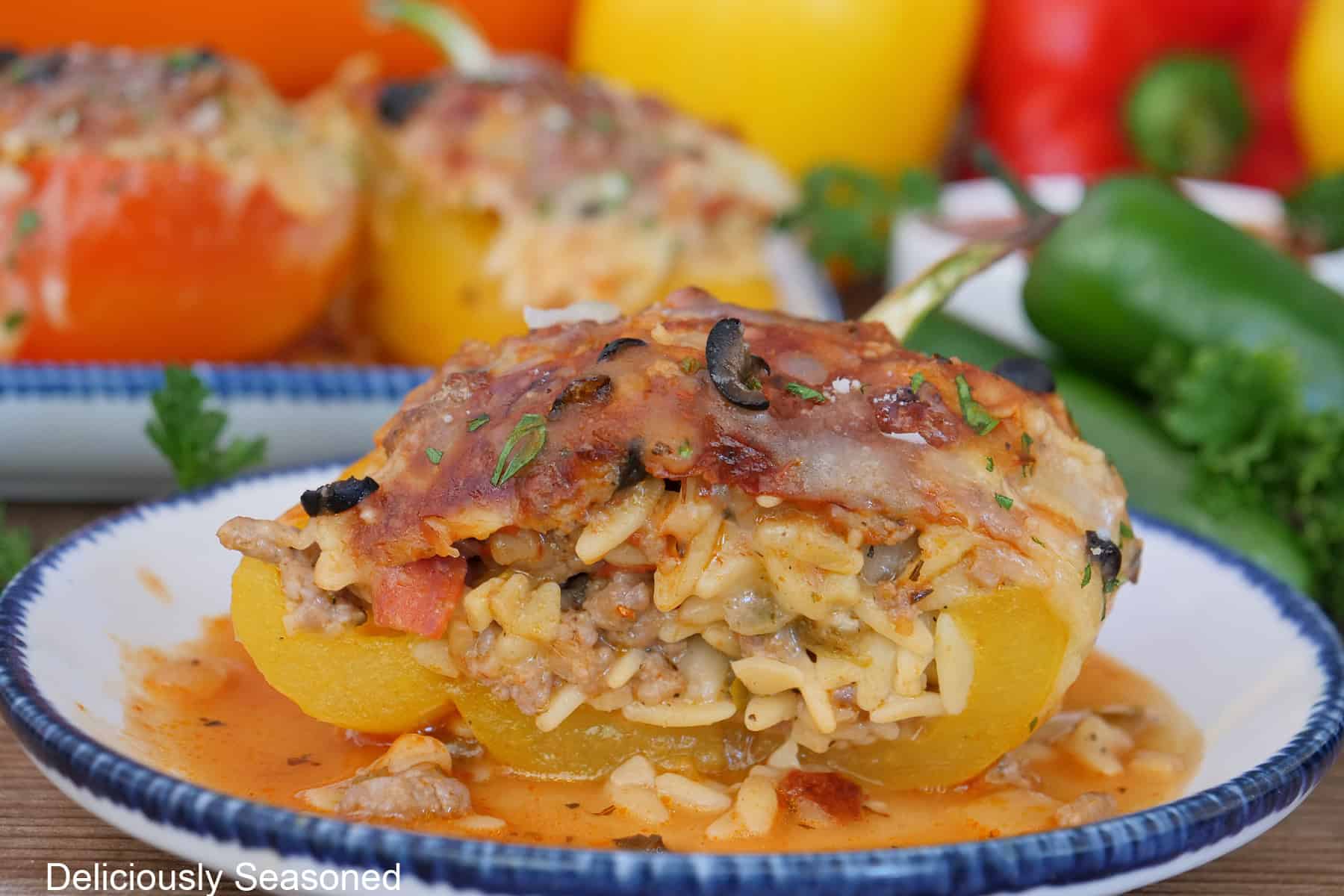 A white plate with blue trim with a yellow bell pepper stuffed with pepperoni pizza ingredients, orzo, topped with melted cheese, with red and yellow bell peppers in the background.