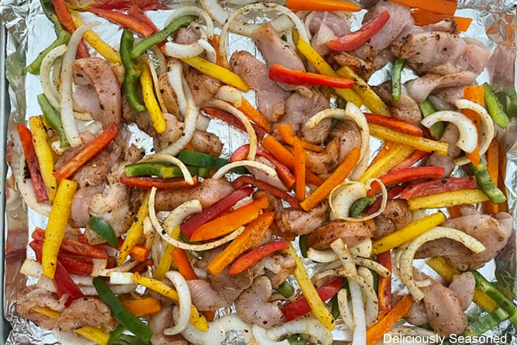 A baking sheet lined with foil with chicken fajitas spread out in an even layer on it.