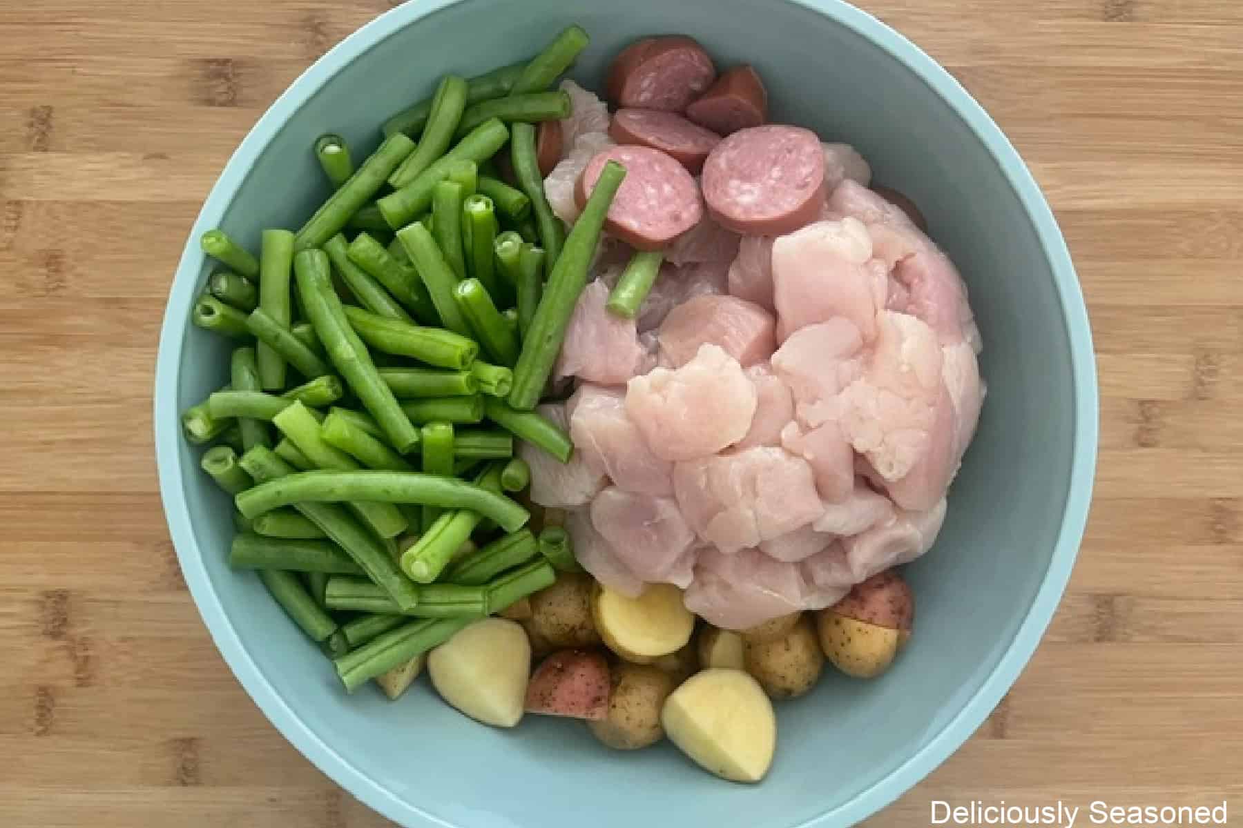 A wood surface with a light blue bowl filled with diced chicken, sliced sausage, fresh green beans, and potatoes.