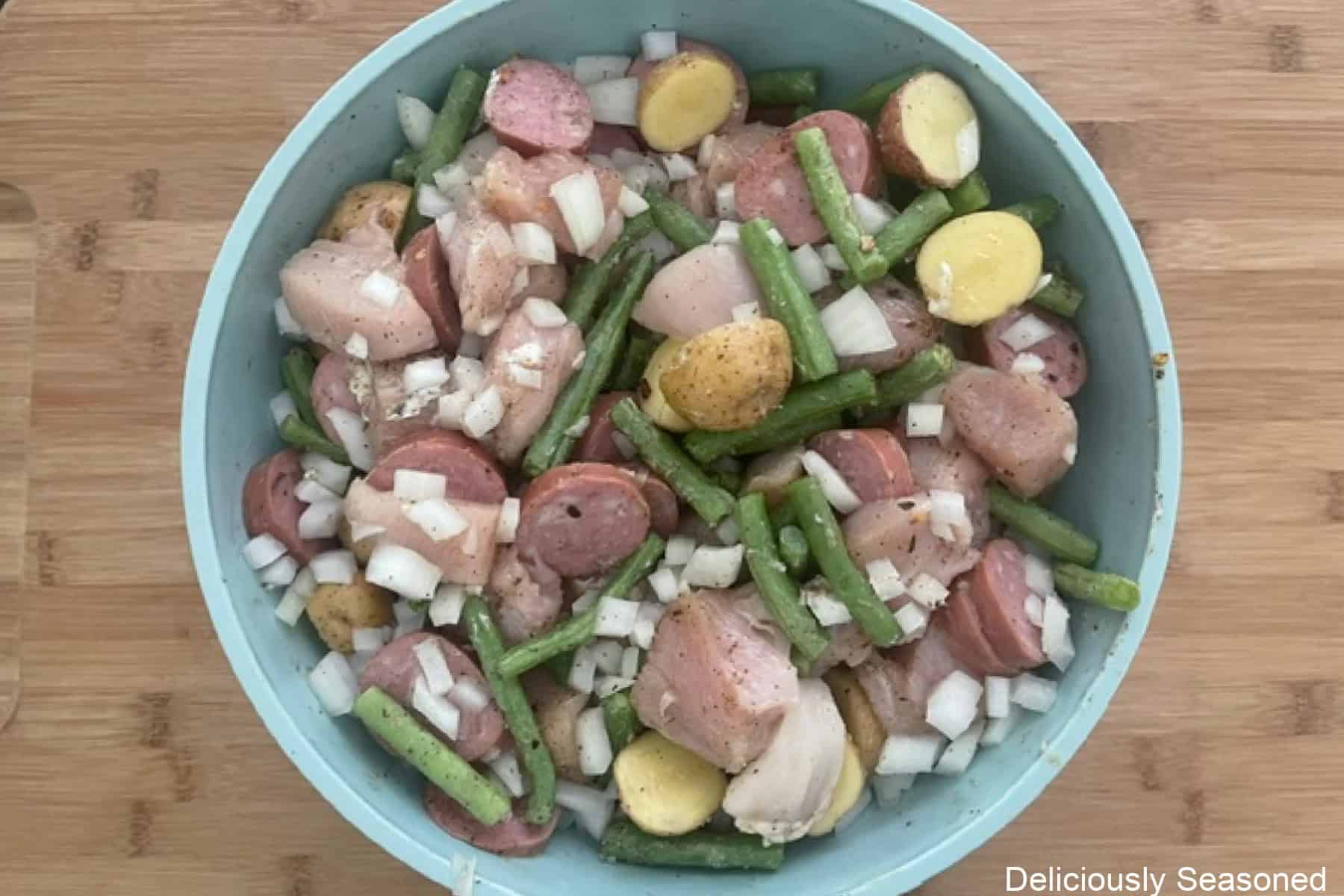 A wood surface with a light blue bowl filled with all the ingredients to make sheet pan chicken and sausage all mixed together.