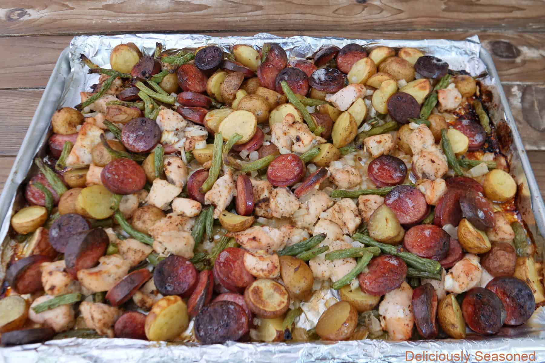 A foil lined baking sheet, placed on a wood surface, with sheet pan chicken and sausage right after being removed from the oven.