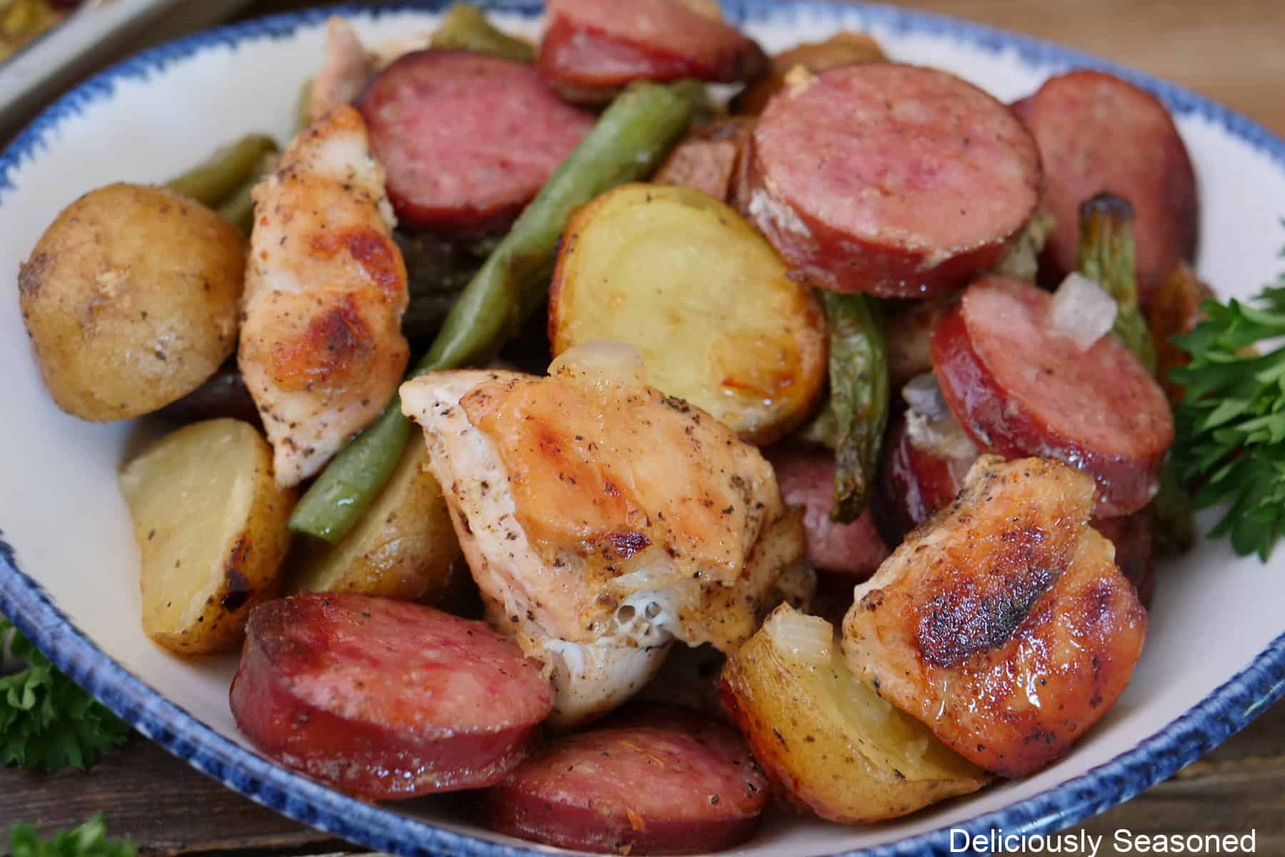 A serving of sheet pan chicken, sausage, potatoes, and green beans in a white bowl with blue trim.