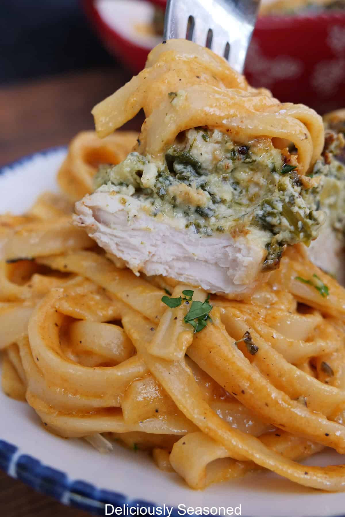 A bite of spinach artichoke stuffed chicken on a fork held above a bowl full of creamy fettuccine pasta in a red pepper sauce.