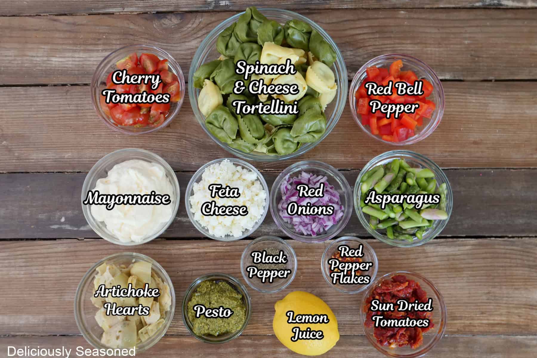 A rustic wood surface with all the ingredients placed in glass bowls with the ingredients to make tortellini pasta salad placed on it.