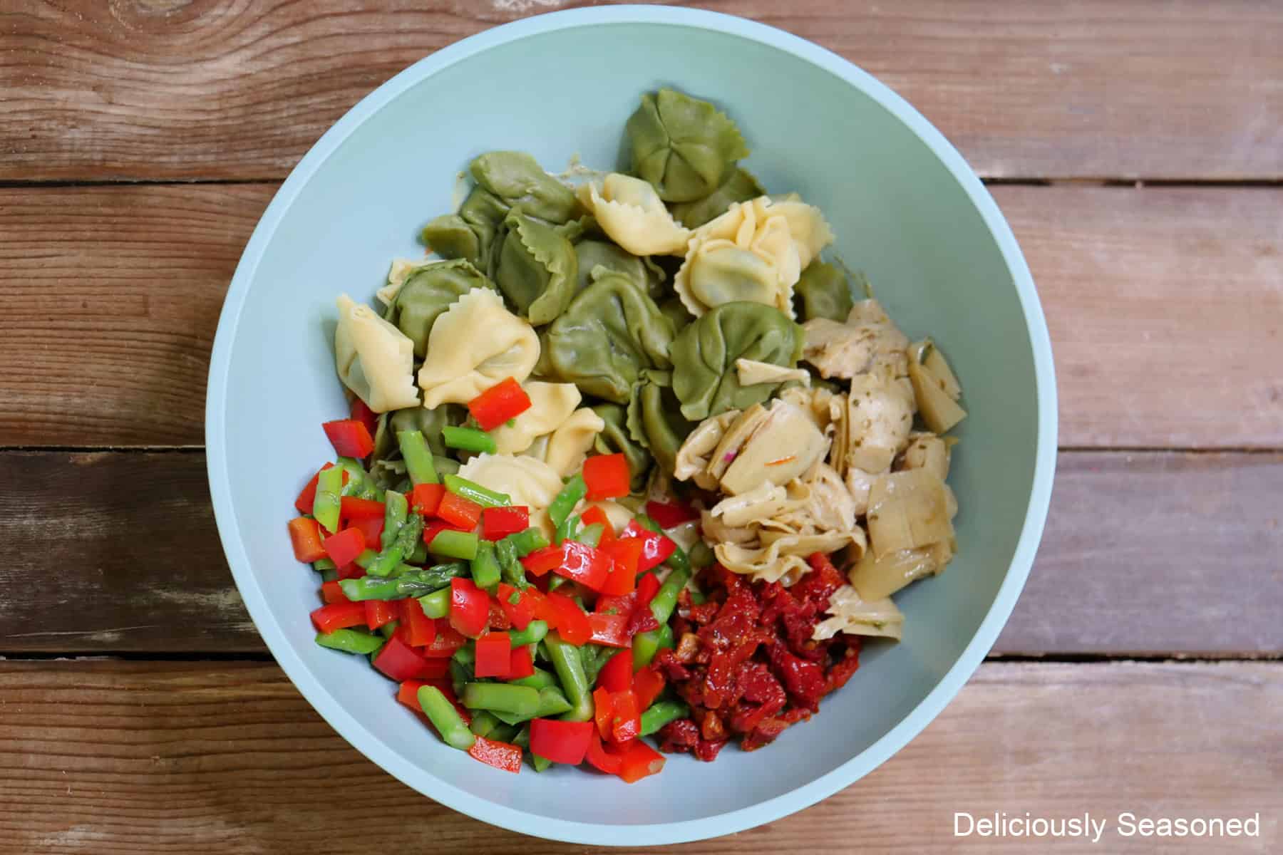 A rustic wood surface with a light blue bowl filled with tortellini pasta salad ingredients in it.