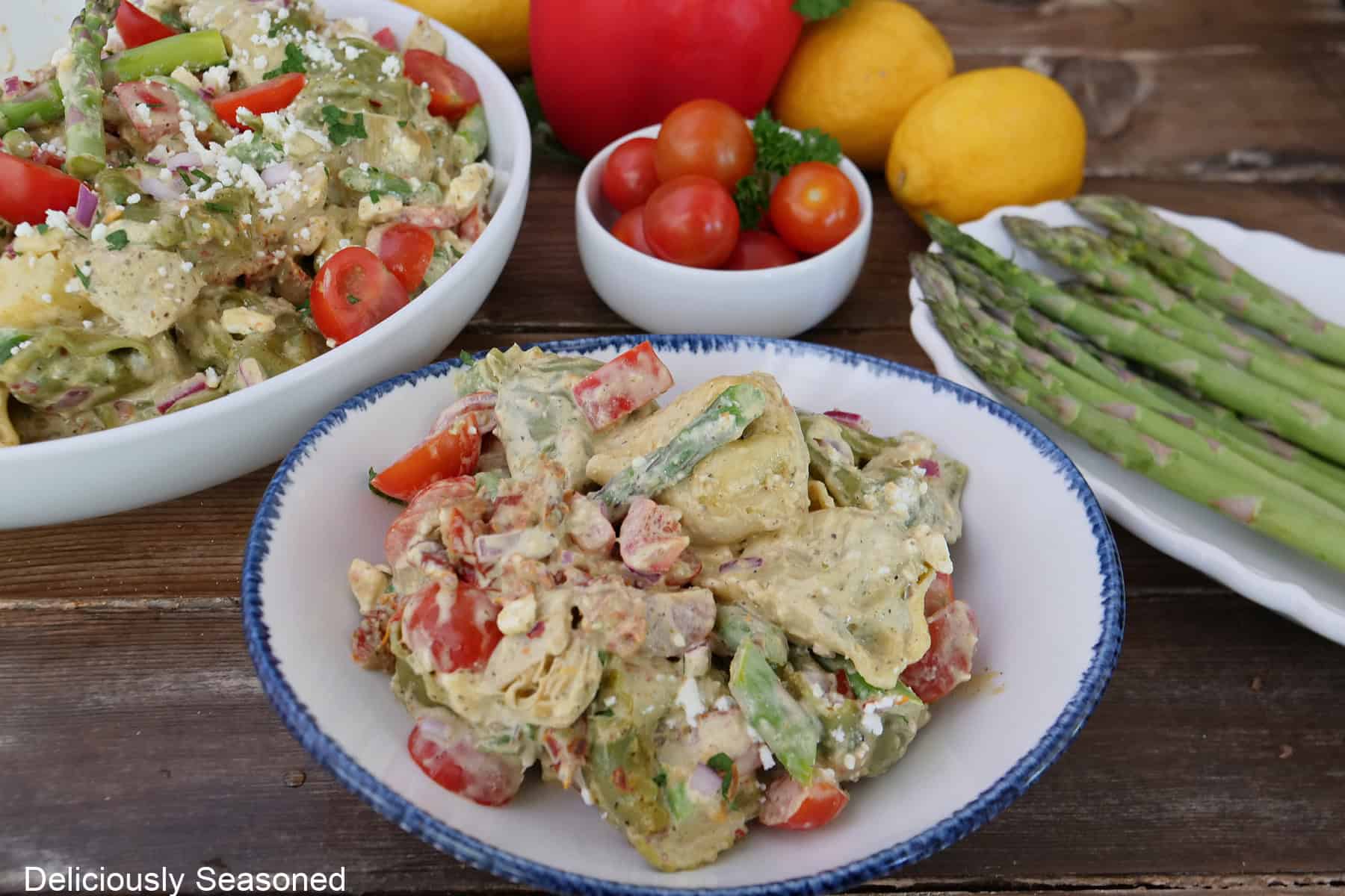 A rustic wood surface with two white bowls filled with tortellini pasta salad with asparagus spears on a white plate and tomatoes in a white bowl in the background.