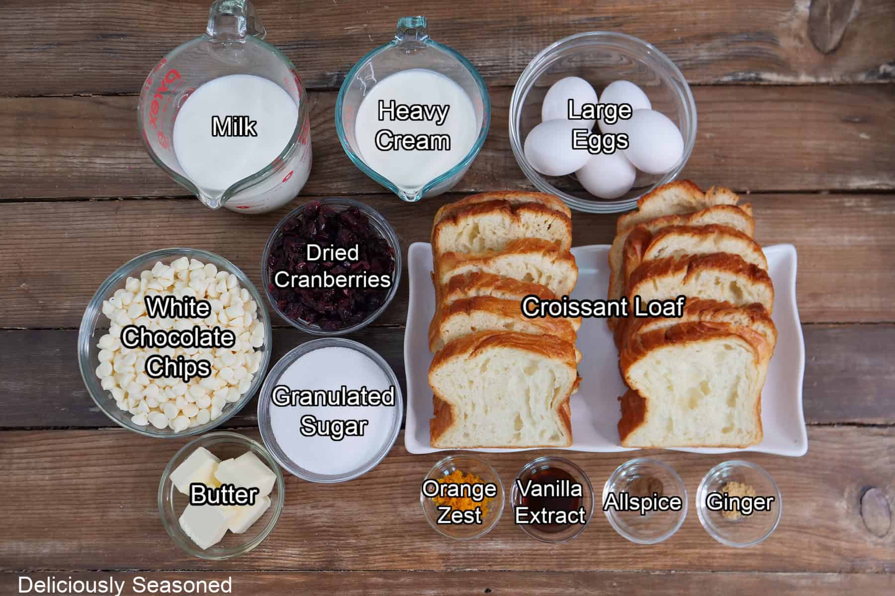 A rustic wood surface with all the ingredients placed on it to make white chocolate cranberry bread pudding.