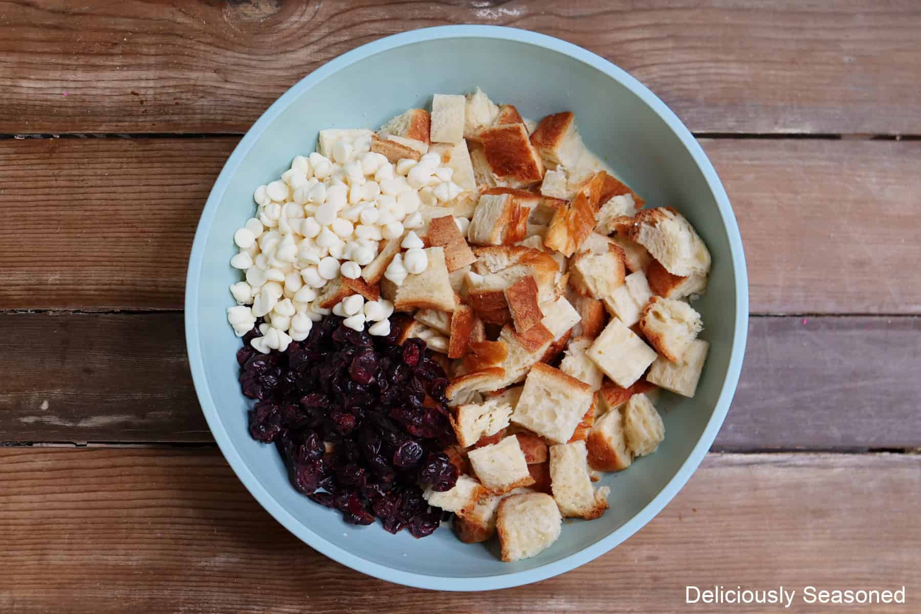 A wood surface with a light blue bowl placed on it with bread pieces, cranberries, white chocolate chips in it.