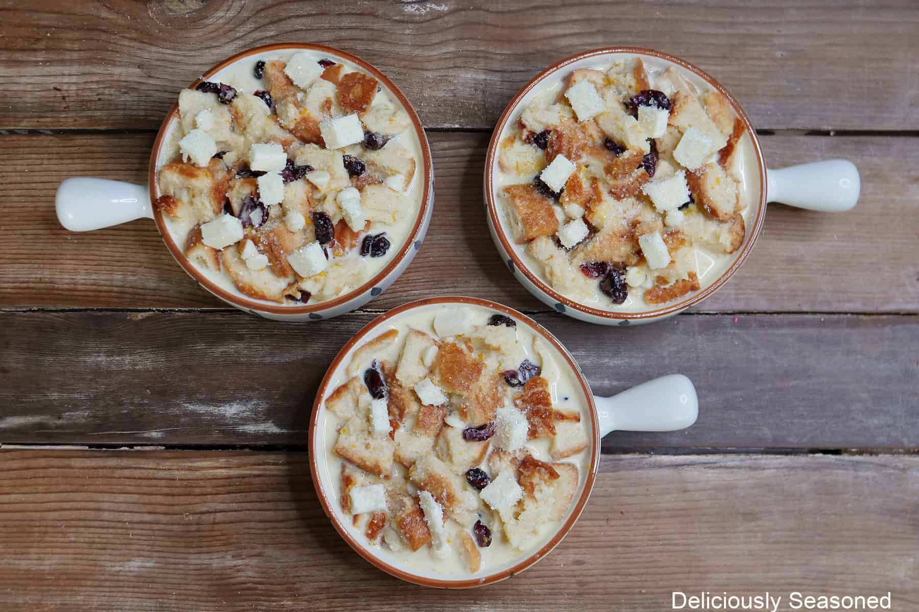 A wood surface with three baking dishes filled with white chocolate cranberry bread pudding in it before going in the oven.