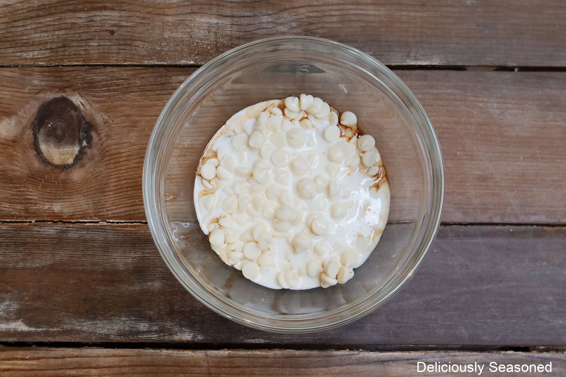 A glass bowl on a wood surface with melting white chocolate chips, vanilla and heavy cream in it.