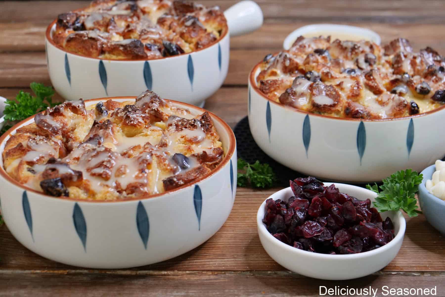 A rustic wood surface with three white and blue baking dishes with white chocolate cranberry bread pudding in them after being baked and drizzled with glaze.