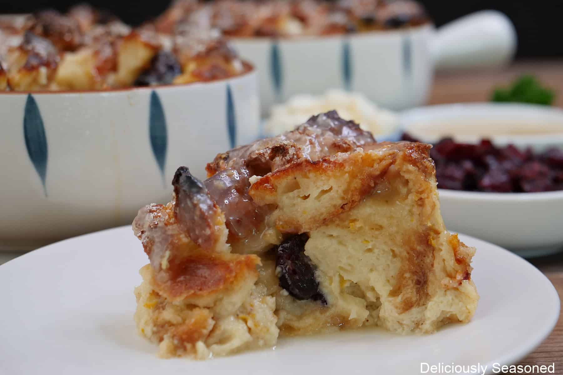 A white plate with a serving of bread pudding on it with two round baking dishes in the background.