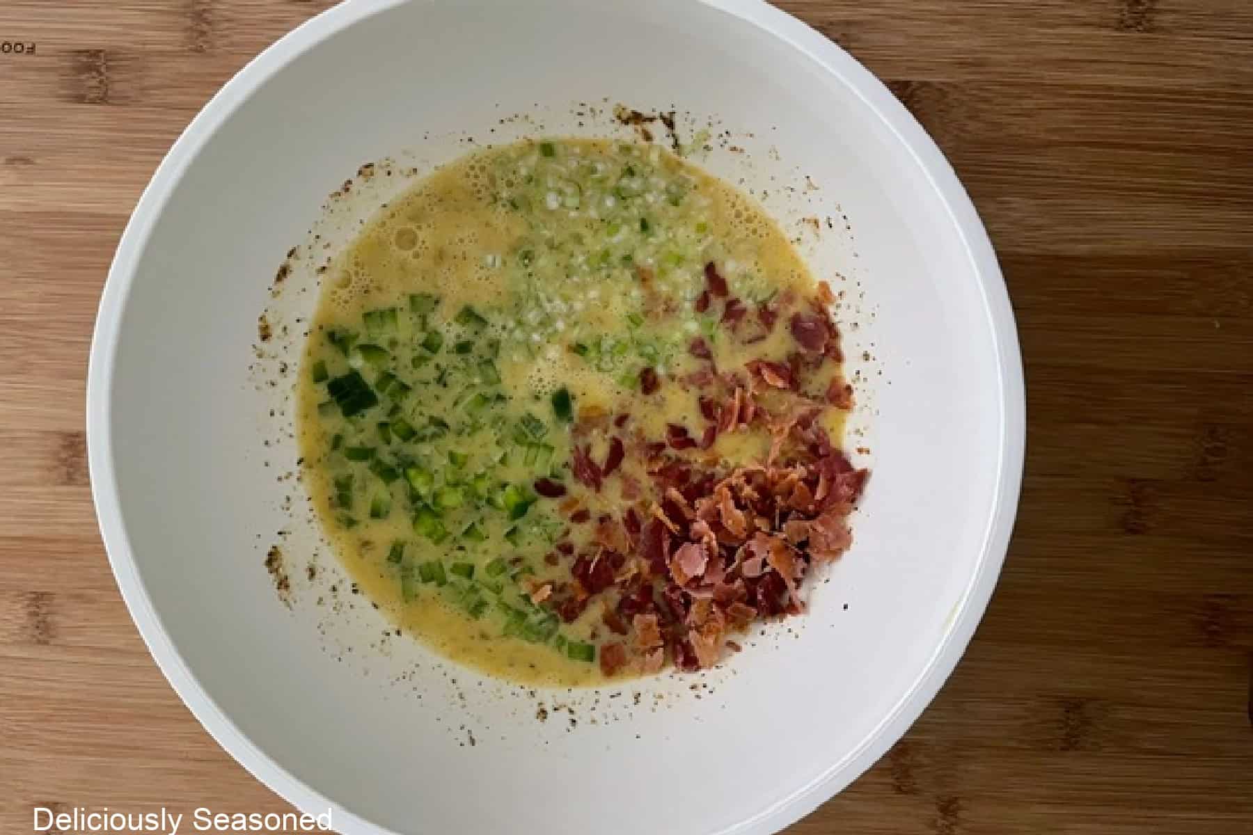 A white bowl with egg mixture along with crumbled bacon, diced onions, and diced bell peppers added to the bowl.