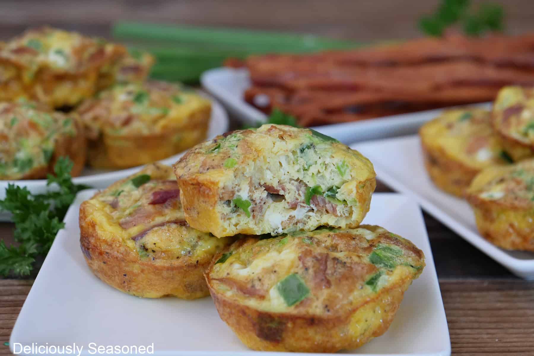 A white square plate with three bacon egg bites on it with a bite taken out of the egg bite on top, with more egg bites and strips of cooked bacon in the background.