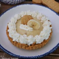 A round white plate with blue trim with a peanut butter cookie crust filled with banana pudding and topped with whipped cream and decorated with a piece of cookie and a slice of banana on top.