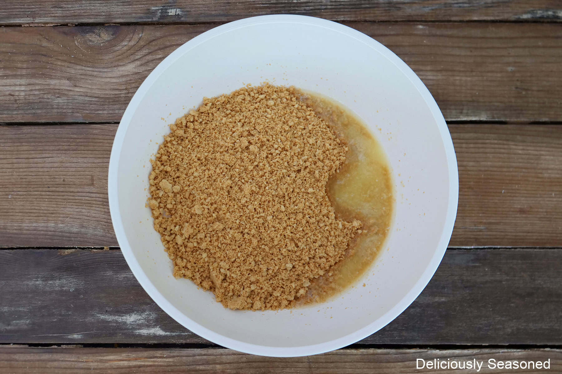 A rustic wood surface with a white mixing bowl filled with crushed peanut butter cookies and melted butter in it.