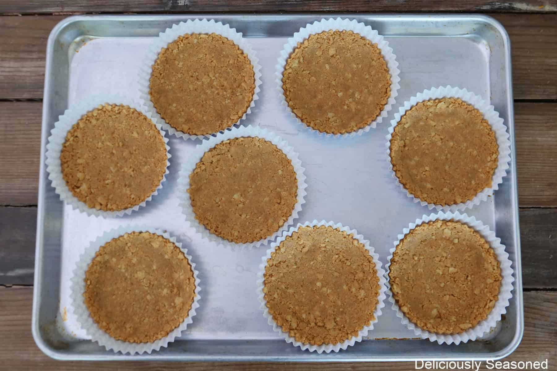 A silver baking sheet with eight 5-inch baking liners filled with crushed peanut butter cookie mixture before being baked in the oven.