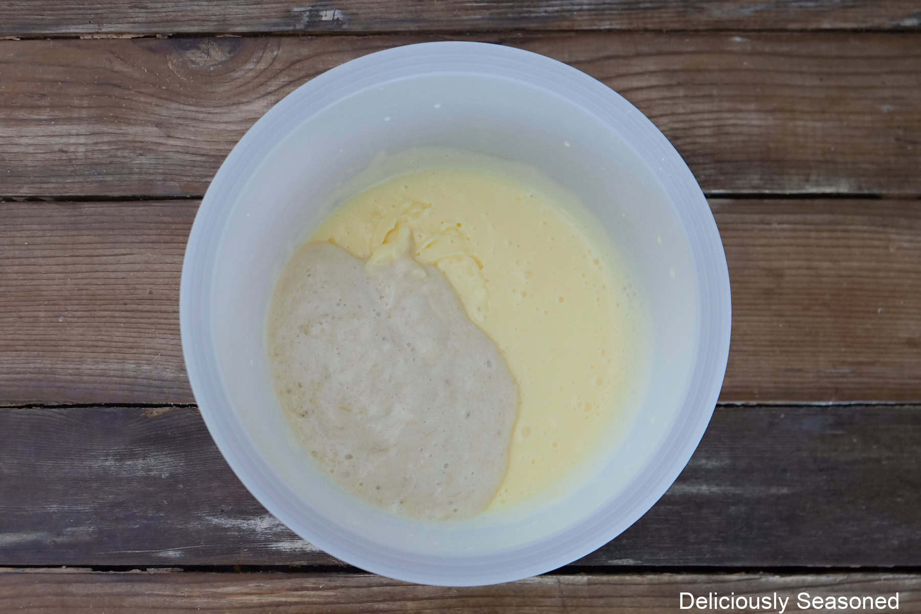 A rustic wood surface with a white mixing bowl with banana pudding mix, milk, and smashed bananas in it.