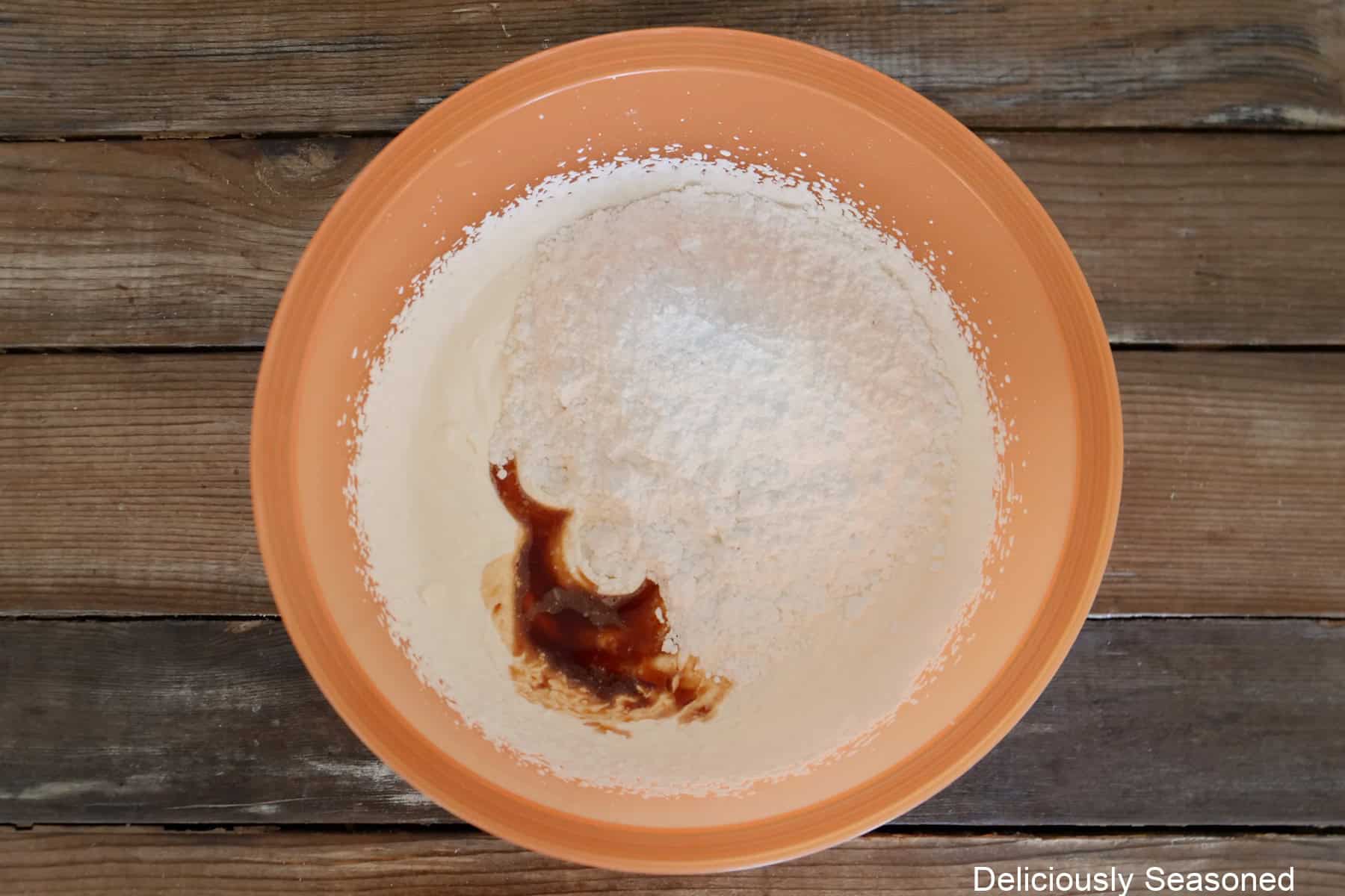 A rustic wood surface with an orange mixing bowl filled with heavy whipping cream, powdered sugar, and vanilla before being mixed all together.