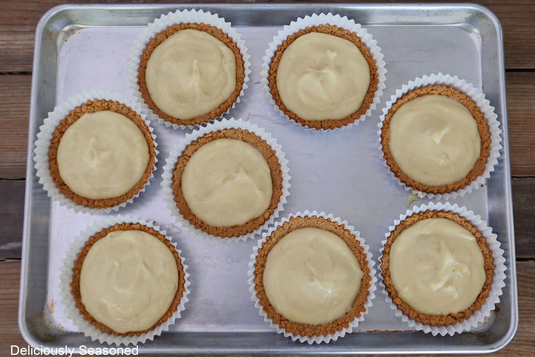 A sliver baking sheet placed on a wood surface with eight 5-inch baking liners filled with banana pudding mixture.