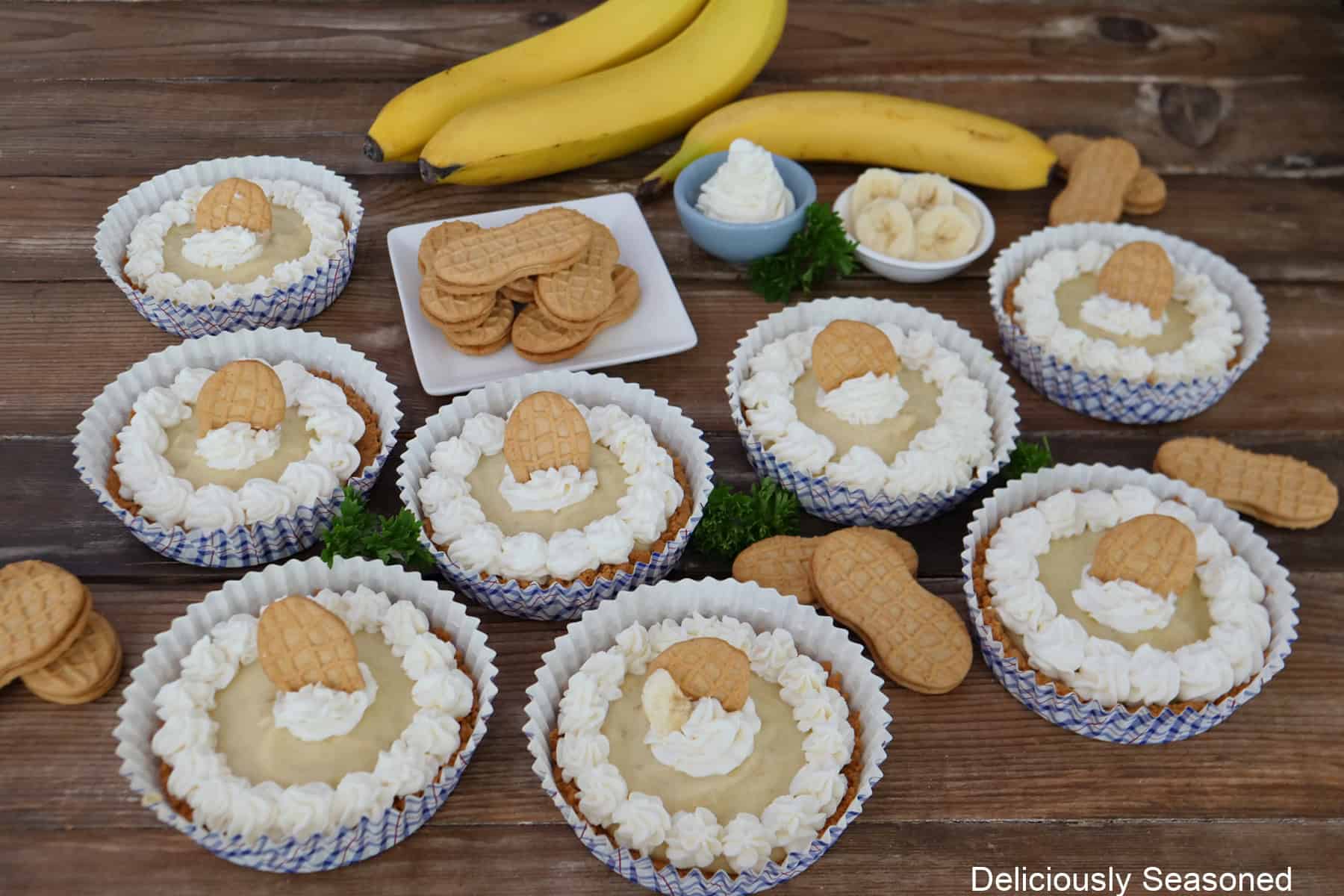A rustic wood surface with eight banana pudding desserts placed on it with three bananas in the background along with Nutter Butter peanut butter cookies randomly placed on the surface.