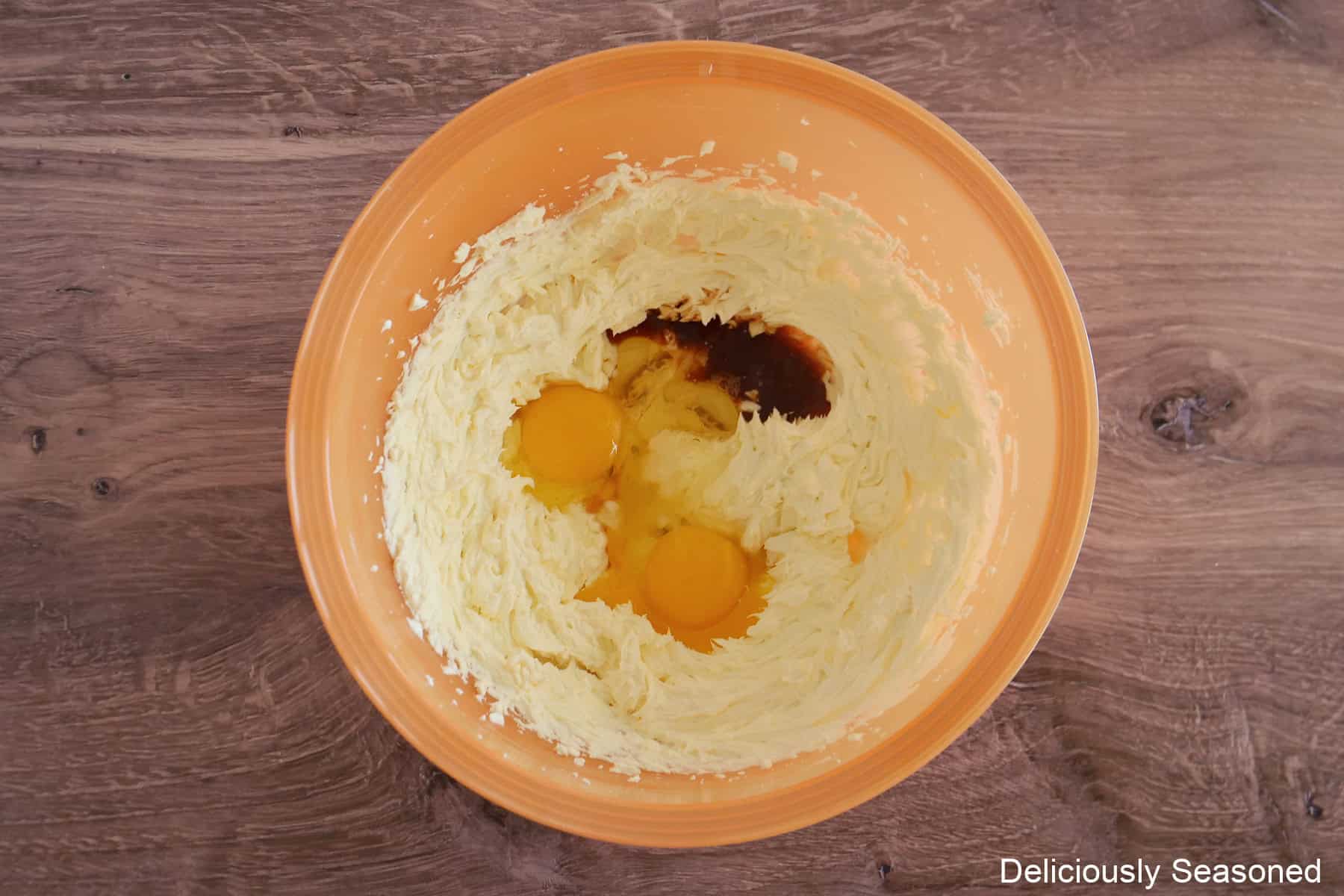 A wood surface with an orange bowl with cheesecake ingredients in it before being all mixed together.
