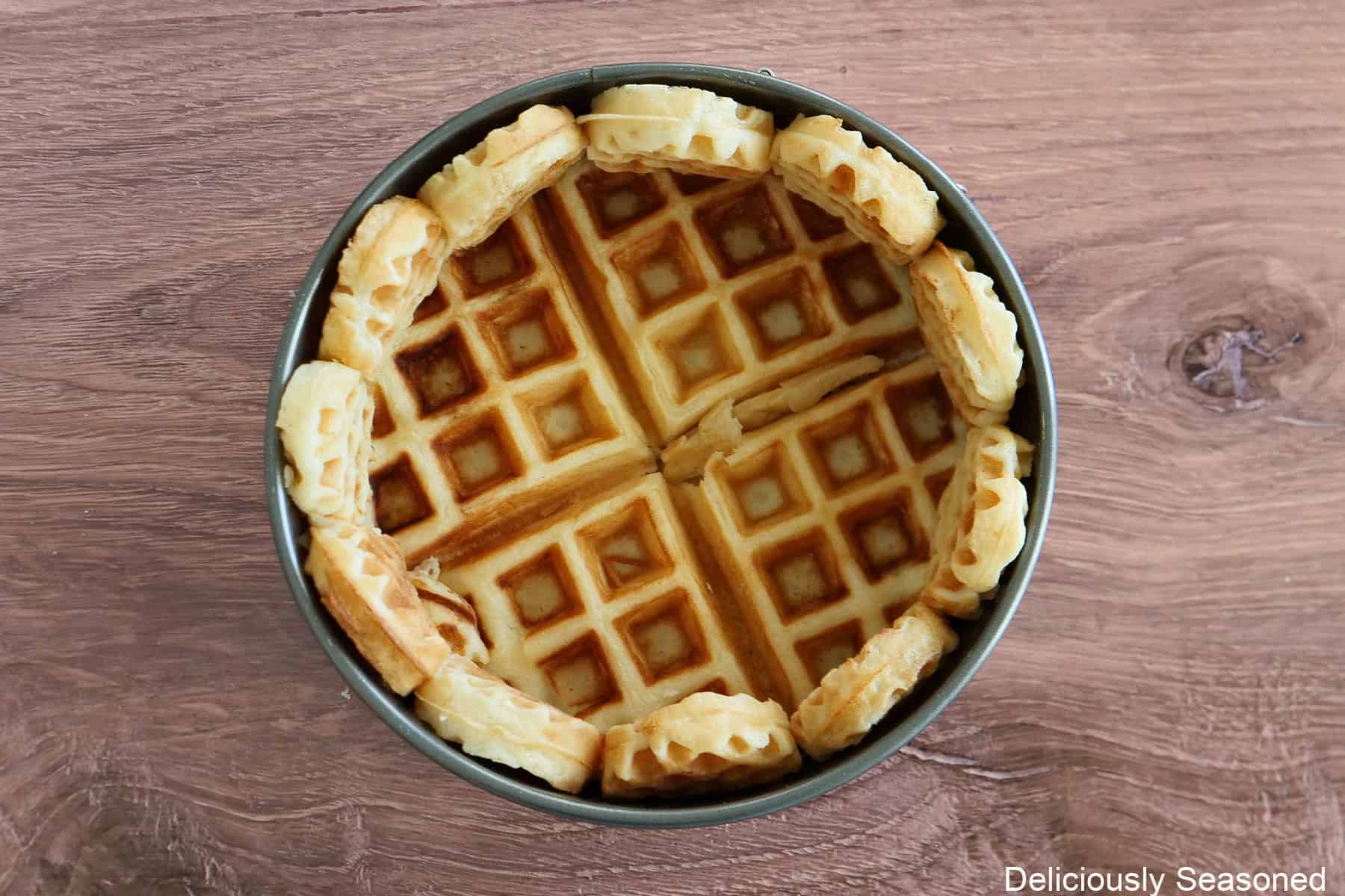A wood surface with a 6 inch springform pan lined with mini waffles along the sides and a big waffle on the bottom.