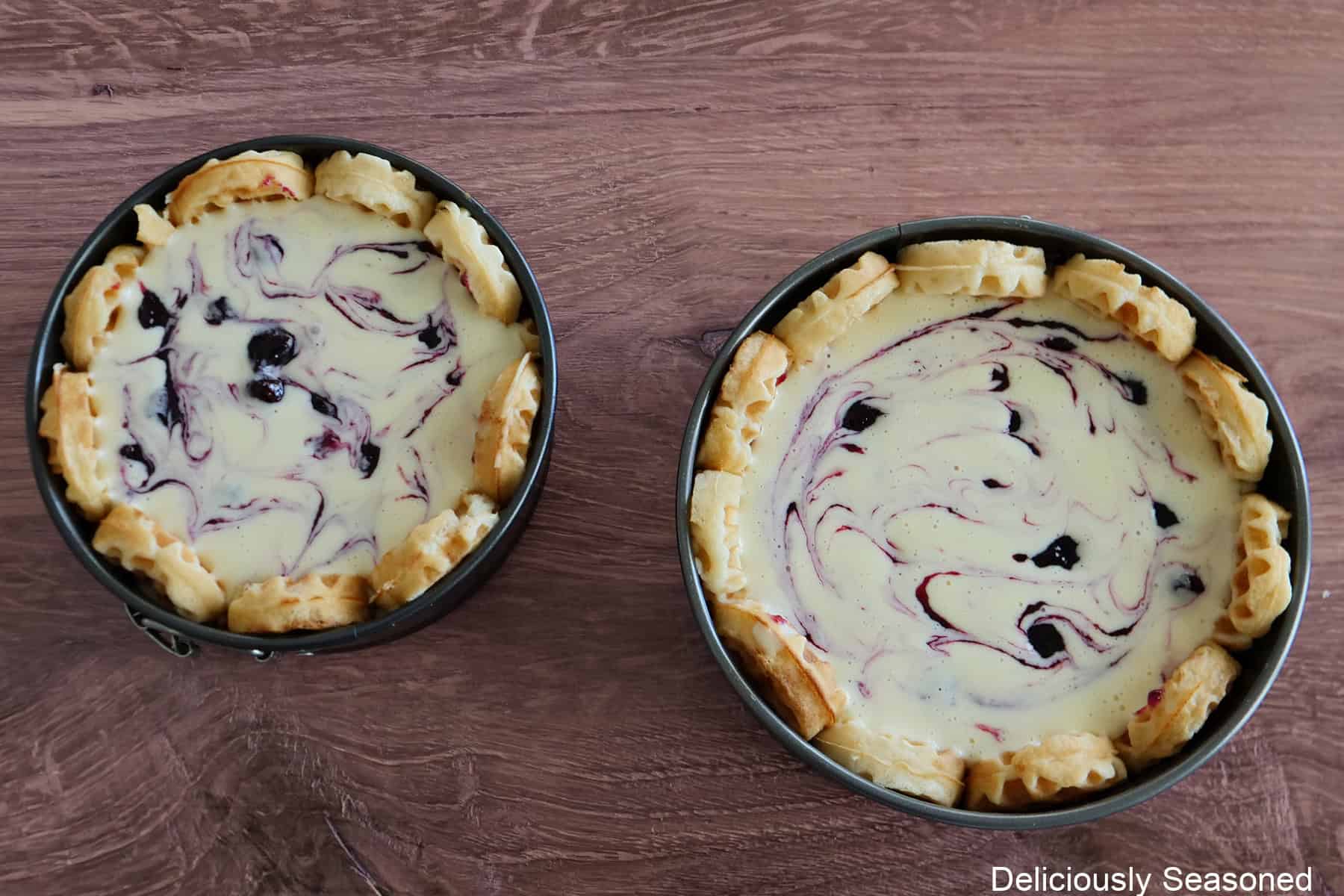 A wood surface with two springform pans filled with blueberry waffle cheesecake before being baked.