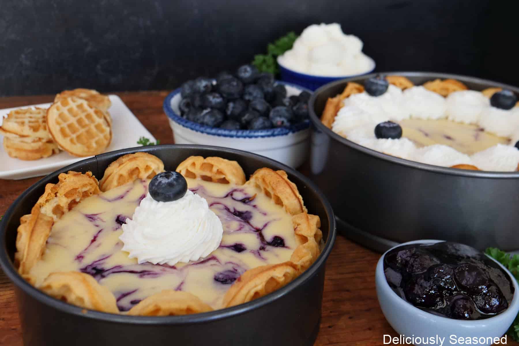 A baked blueberry waffle cheesecake on a wood surface with another cheesecake in the background along with mini waffles and fresh blueberries.