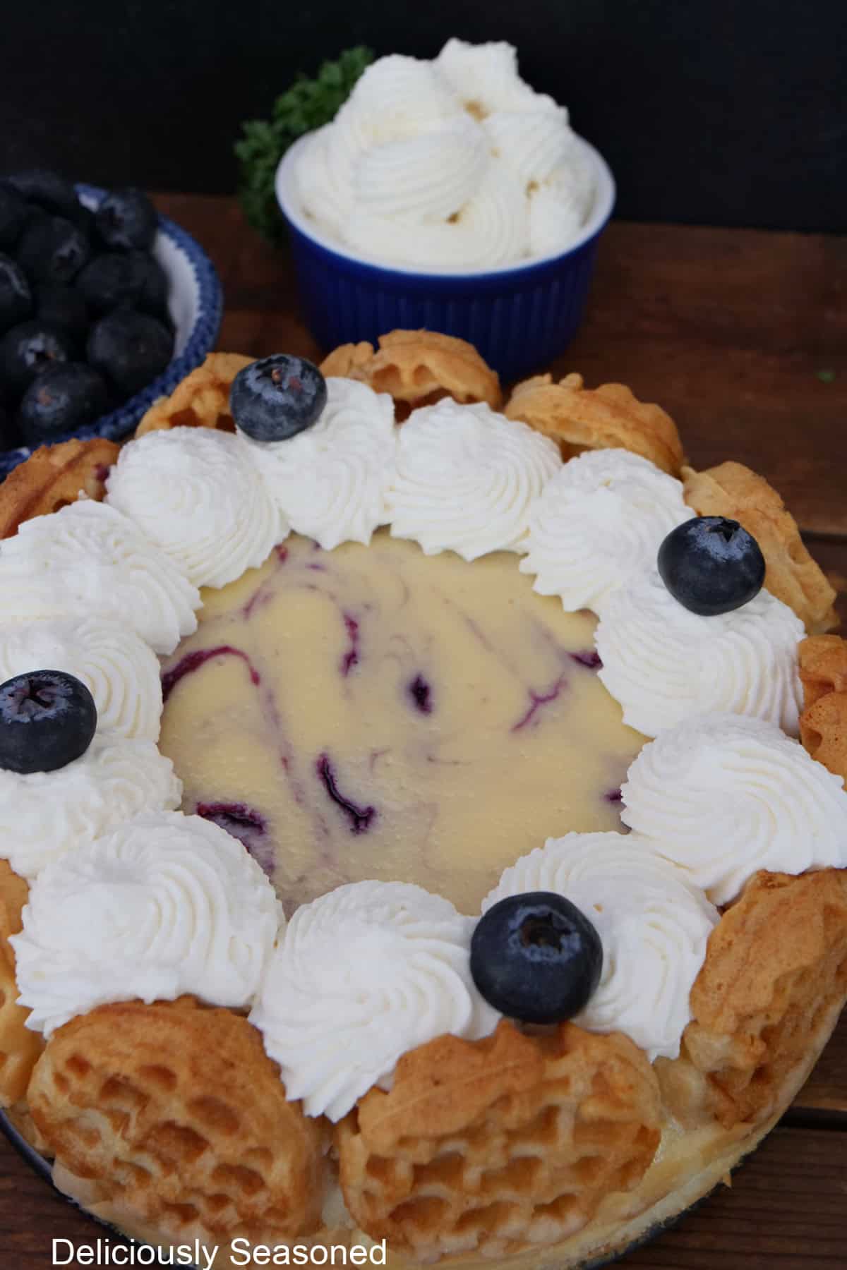 A blueberry waffle cheesecake with two small bowls of whipped cream and blueberries in the background.