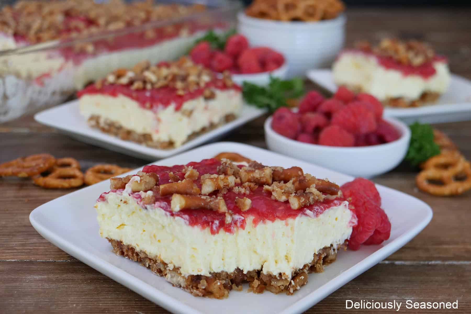 A wood surface with three white plates with a serving of lemon raspberry pretzel salad on them with a small white bowl filled with fresh raspberries.
