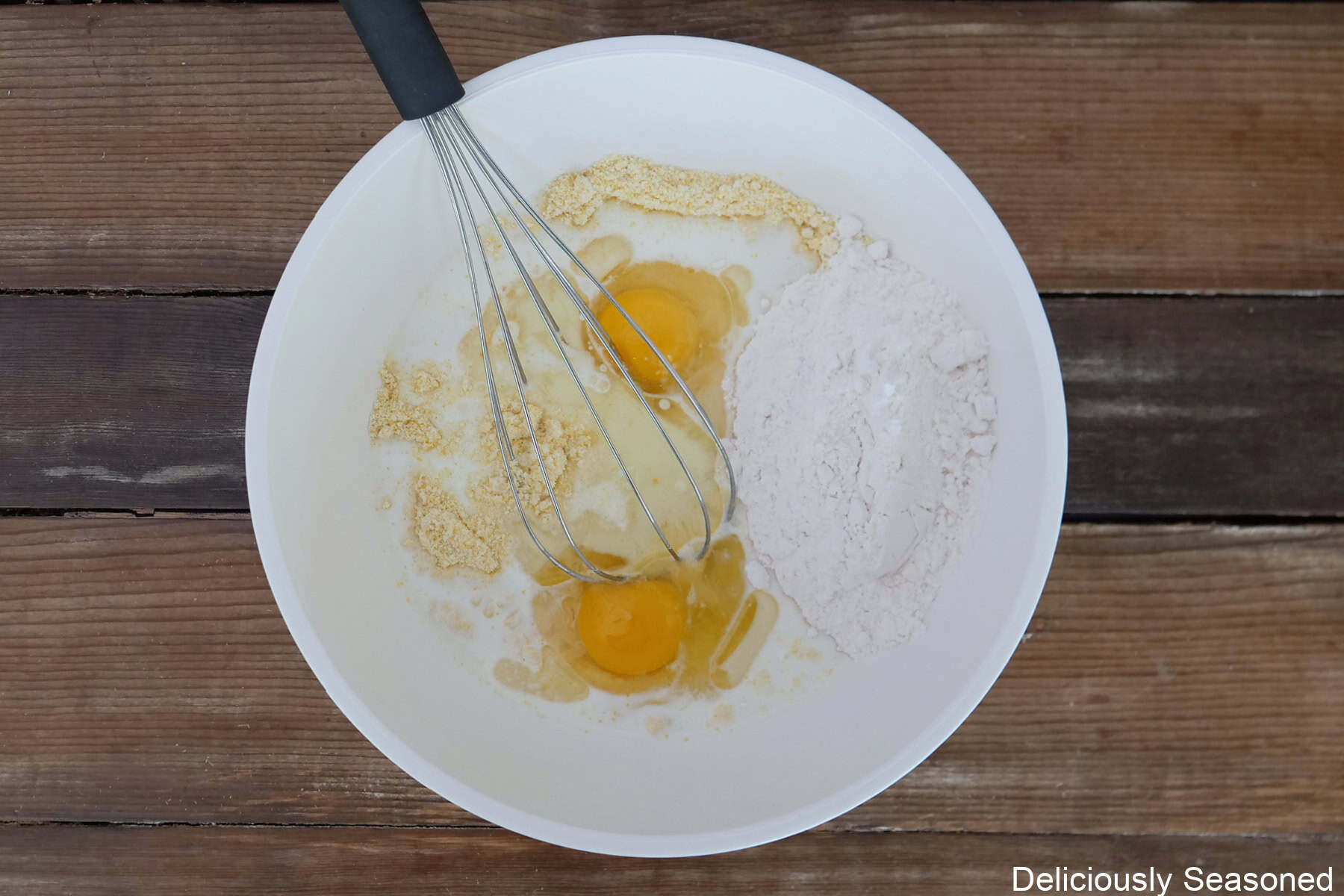 A rustic wood surface with a white mixing bowl filled with cornbread waffle ingredients in it before being mixed together.