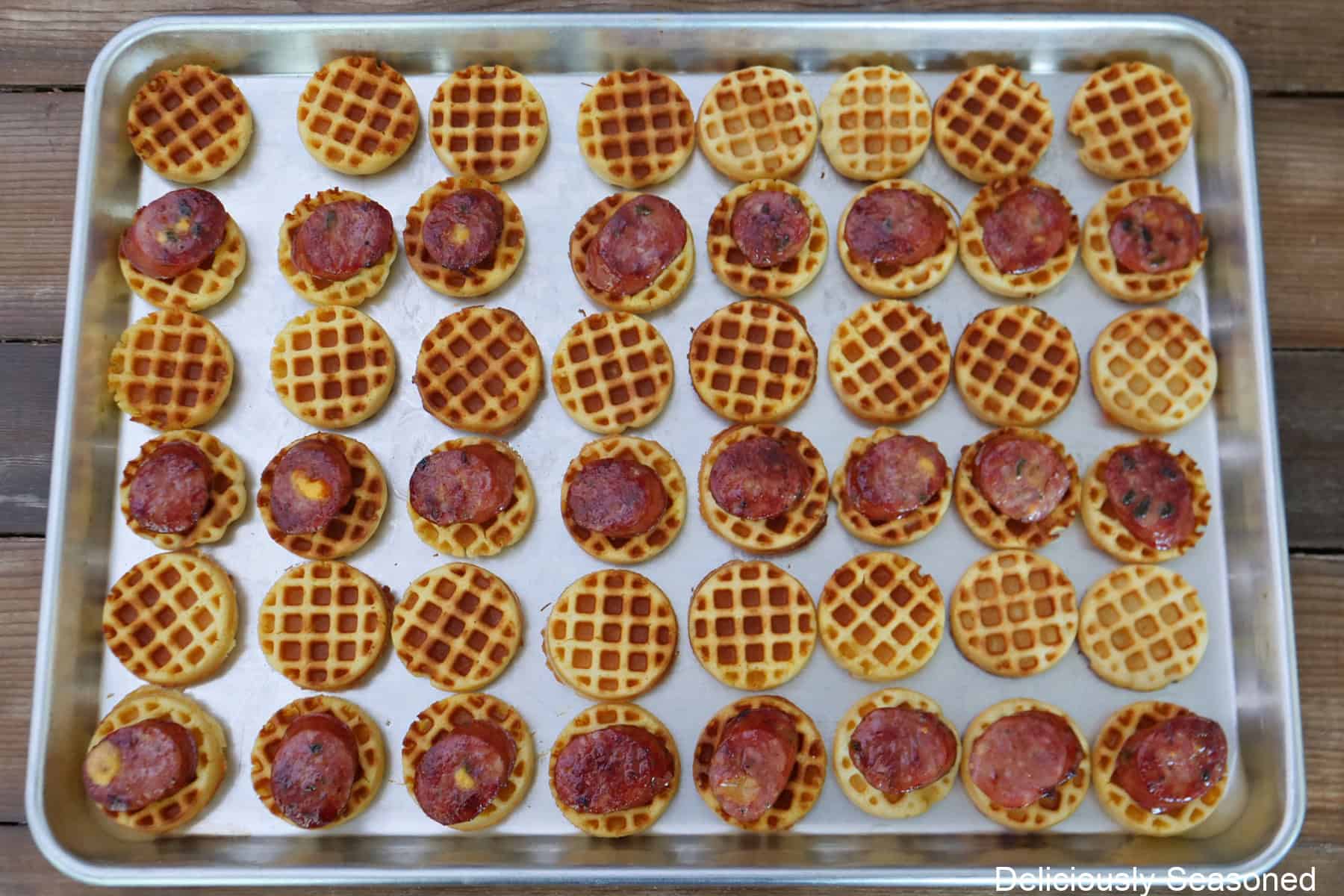 A silver baking sheet with mini cornbread waffles and sausage on them spread out on the baking sheet.