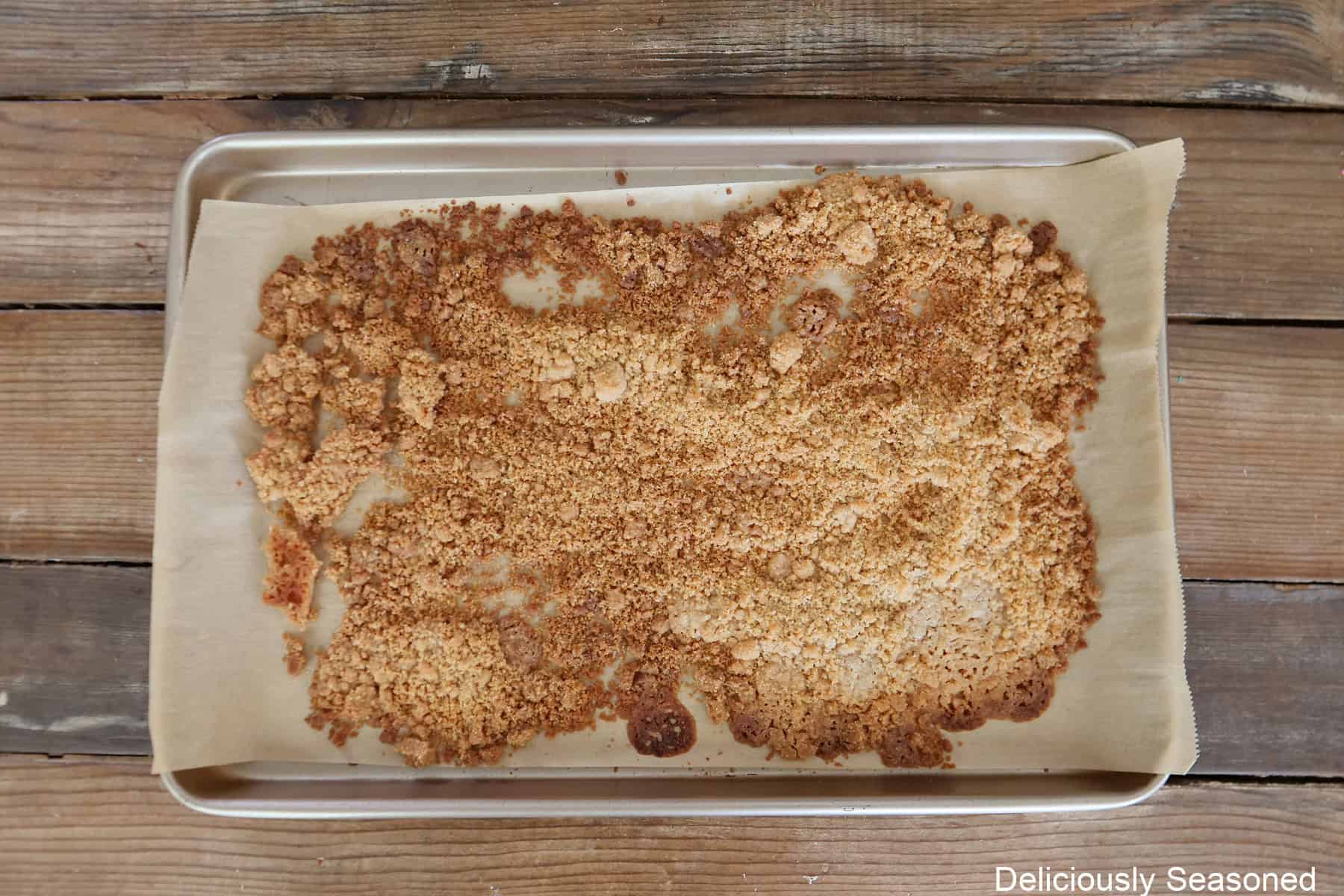 A wood surface with a silver baking sheet with baked streusel topping spread out on parchment paper.