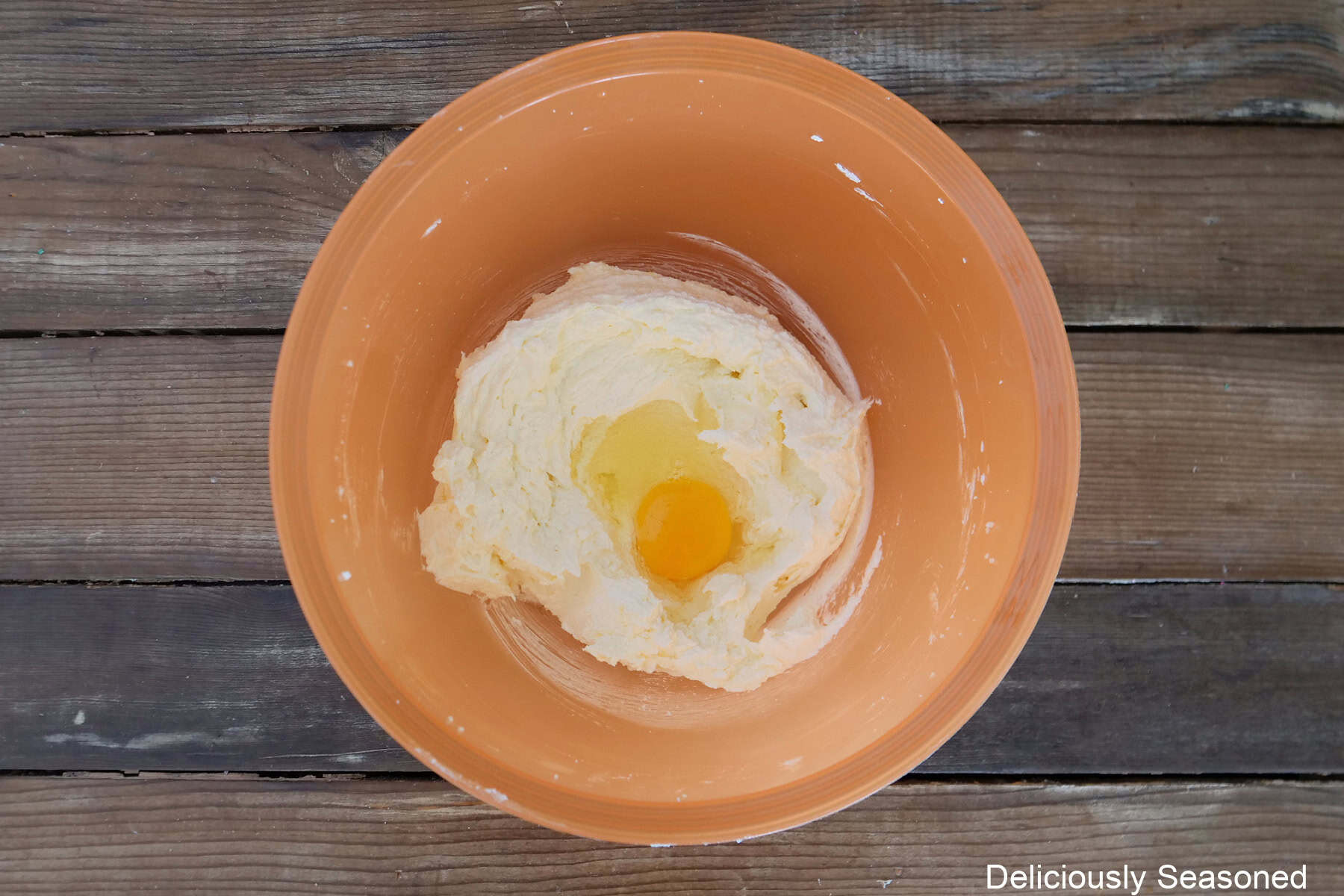 A wood surface with an orange bowl with cupcake batter mixed together and an egg being added to the mixture.