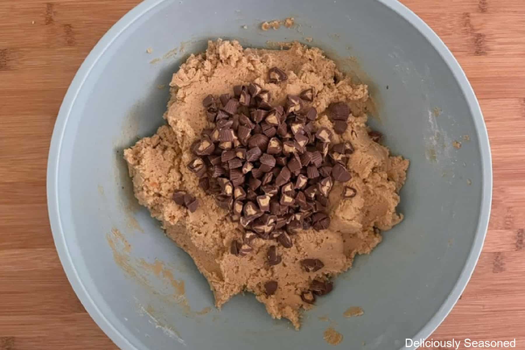 A blue bowl on a wood surface with cookie batter in it and chopped mini peanut butter cups added before being stirred together.