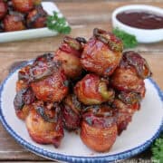 A wood surface with a white plate with blue trim filled with smoked bacon wrapped meatballs stack on it with more in the background and a small white bowl with dipping sauce in the background.