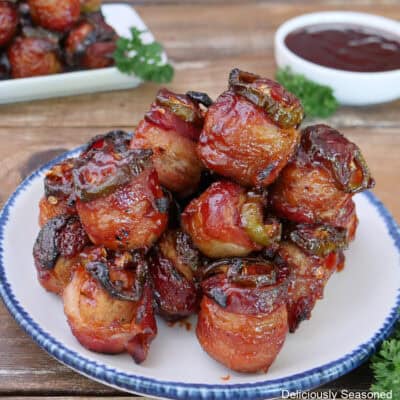 A wood surface with a white plate with blue trim filled with smoked bacon wrapped meatballs stack on it with more in the background and a small white bowl with dipping sauce in the background.