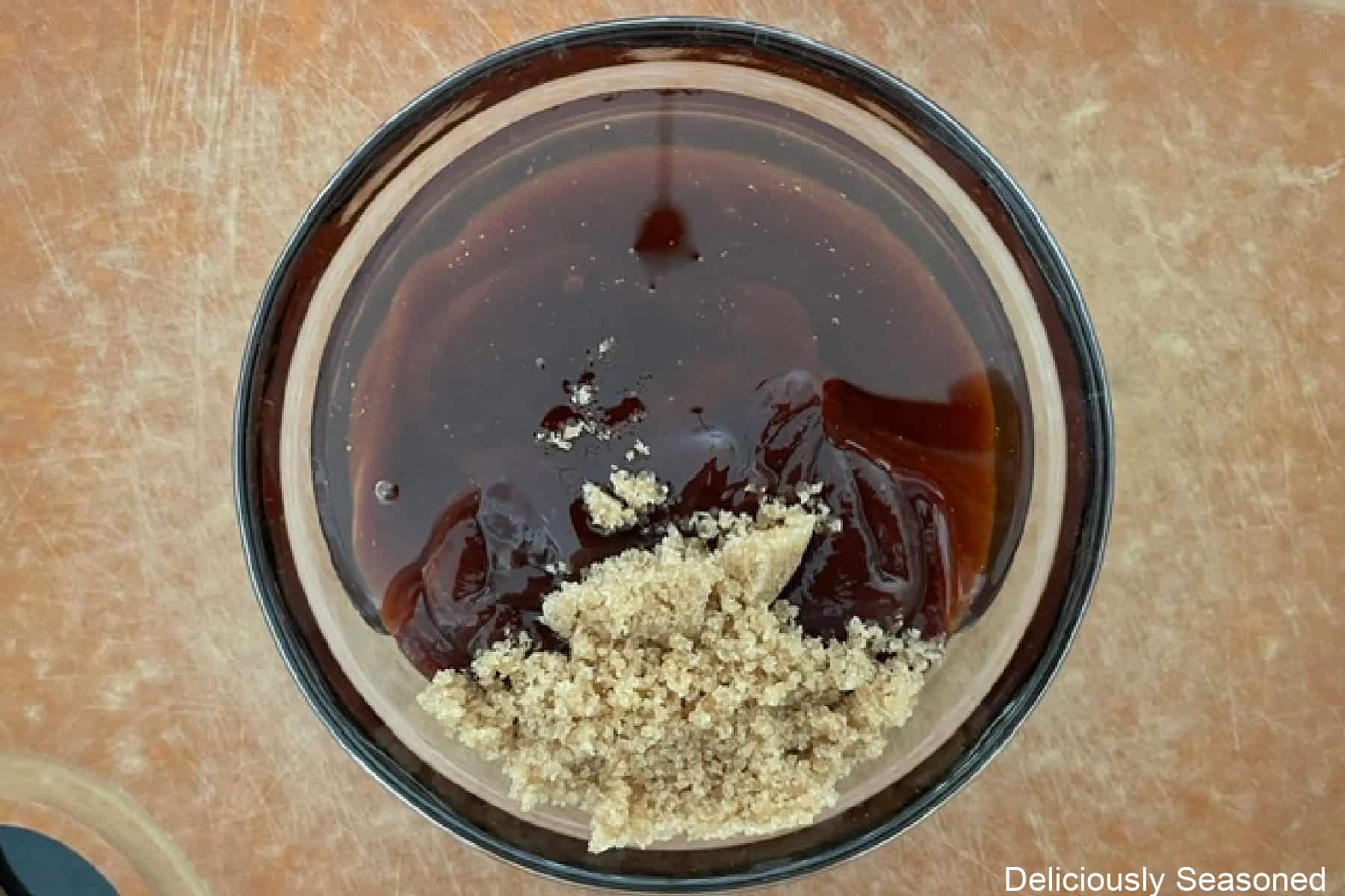 A wood surface with a glass bowl filled with barbecue sauce, honey, and brown sugar before being stir together.