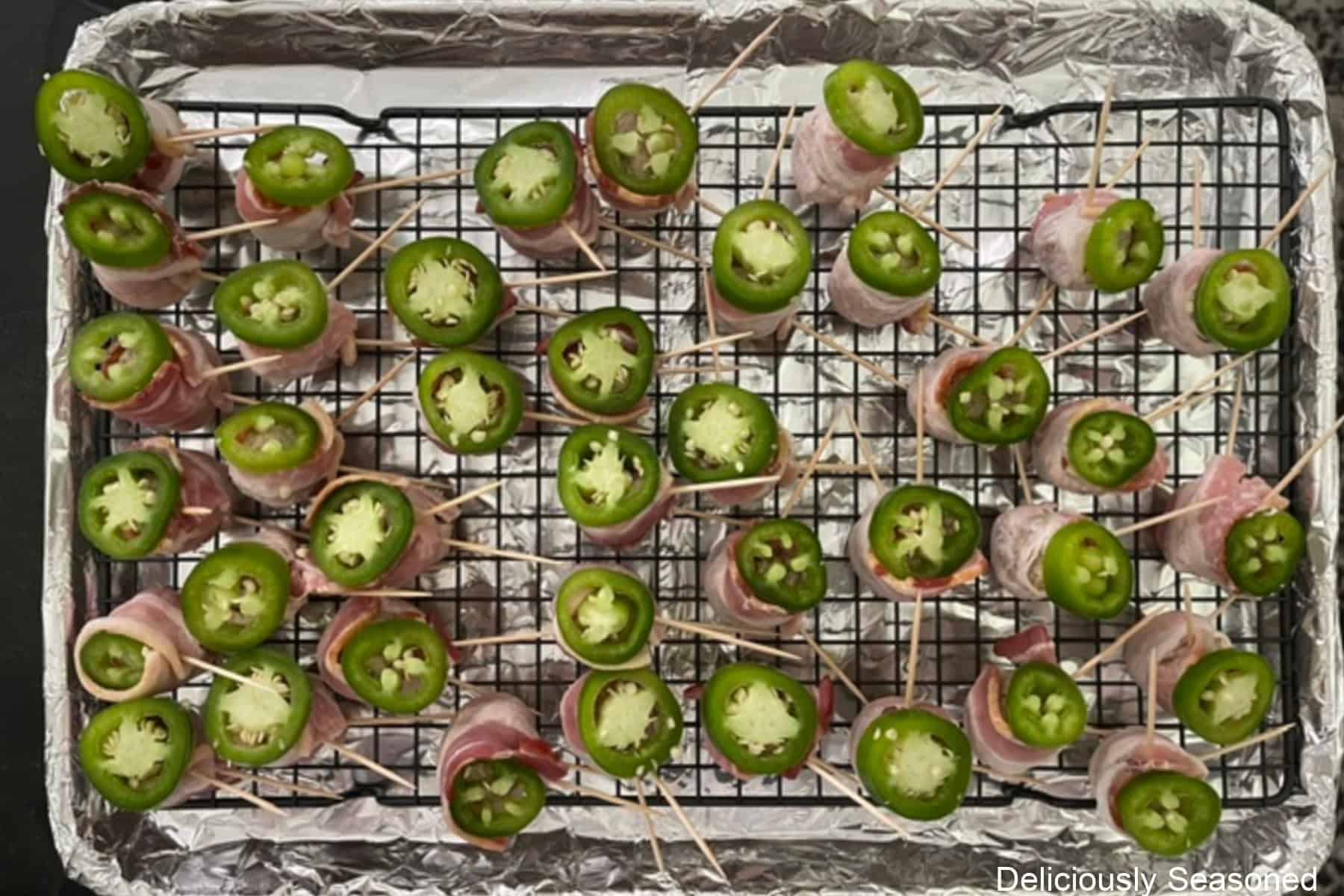 A foil lined baking sheet with a wire rack filled with bacon wrapped meatballs with jalapeno slices on top of the meatballs.