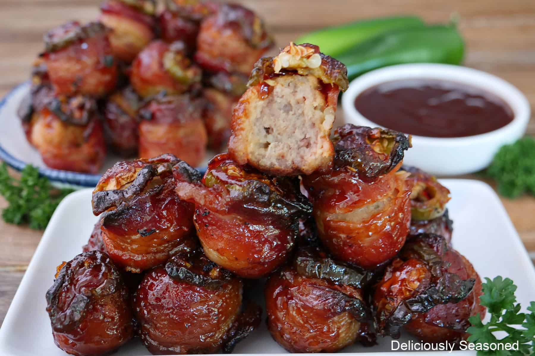 A white plate stacked full of bacon wrapped meatballs with a bite taken out of the meatball on top, and a small white bowl filled with dipping sauce and more meatballs in the background.