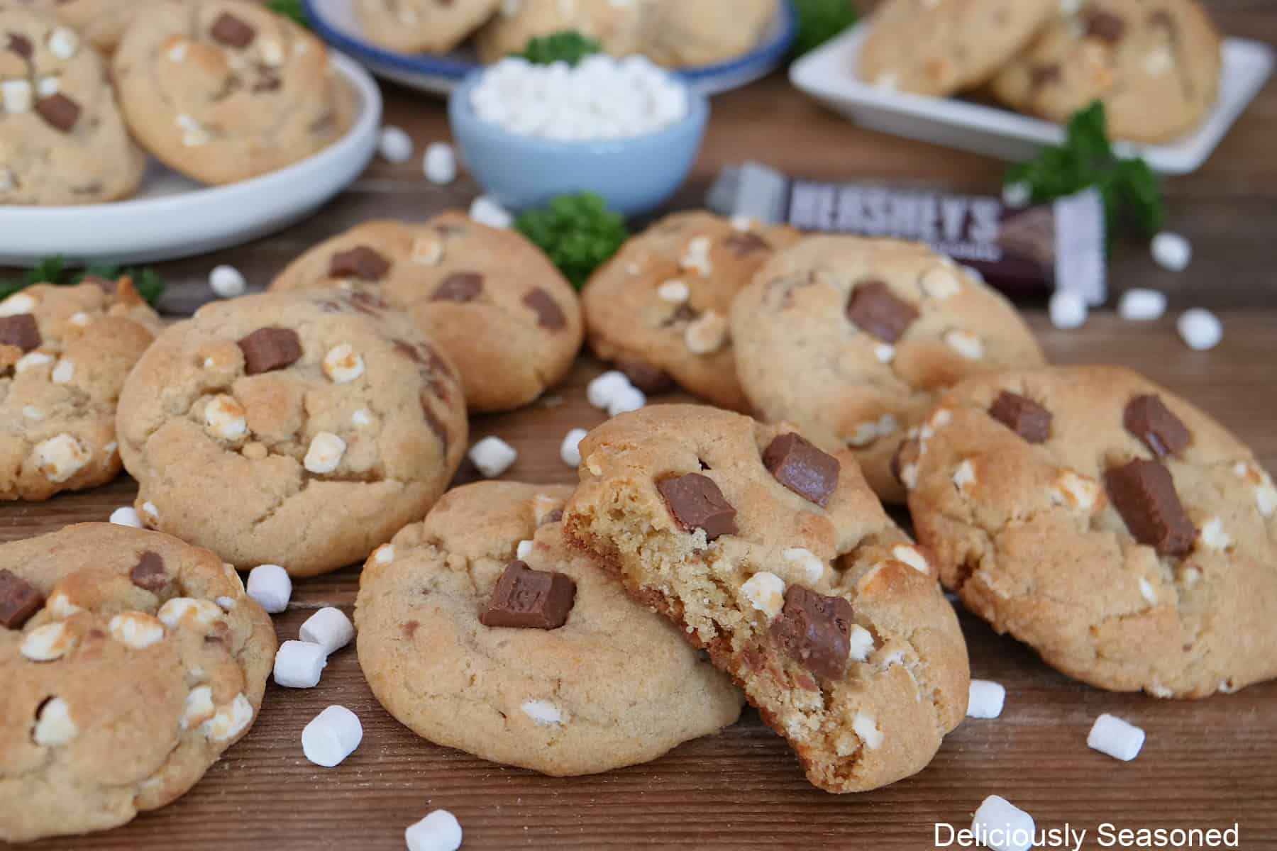 A wood surface with over a dozen s'mores cookies on it with little marshmallow bits randomly scattered around on the surface.