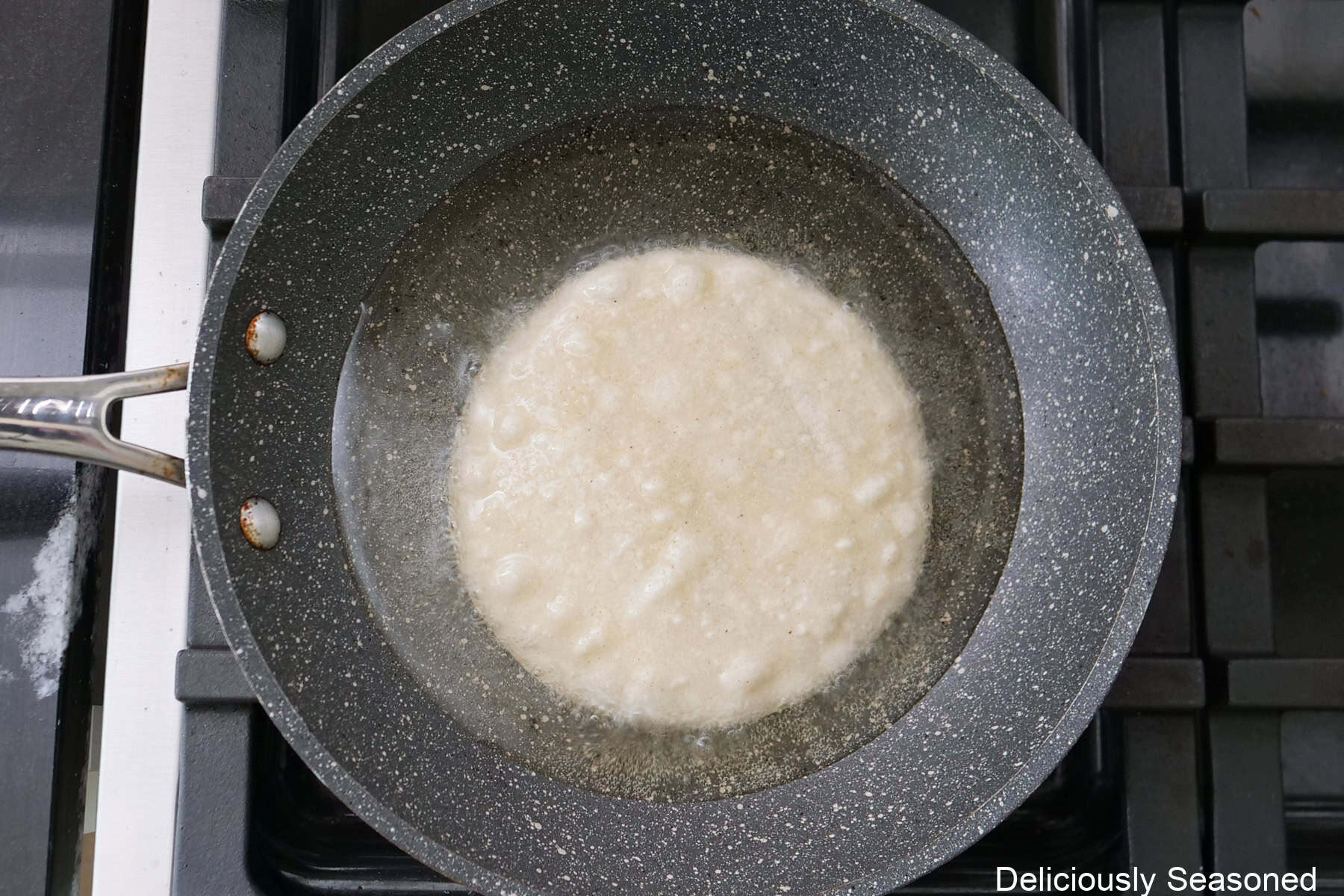 A frying pan on the stovetop with a corn tortilla being fried in oil.