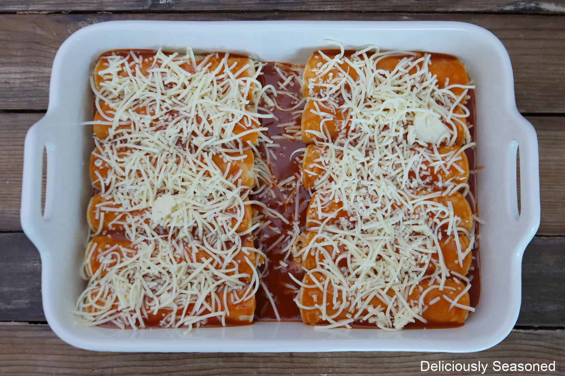 A white 13 x 9 baking dish with ten enchiladas in it topped with Monterey jack cheese, before being baked in the oven.