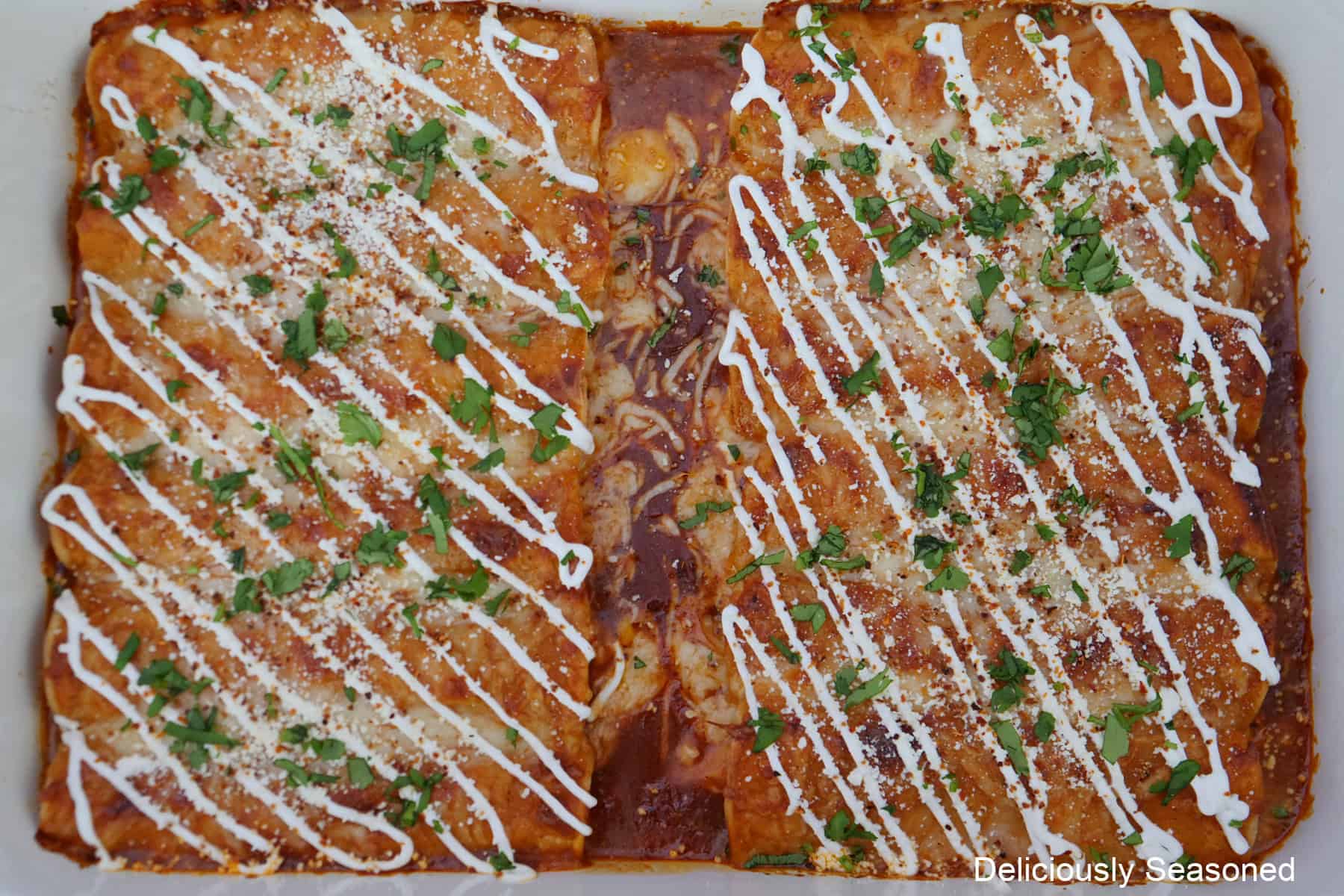 A 13 by 9 baking dish with baked chicken enchiladas in it drizzled with sour cream and topped with Cotija cheese and cilantro.