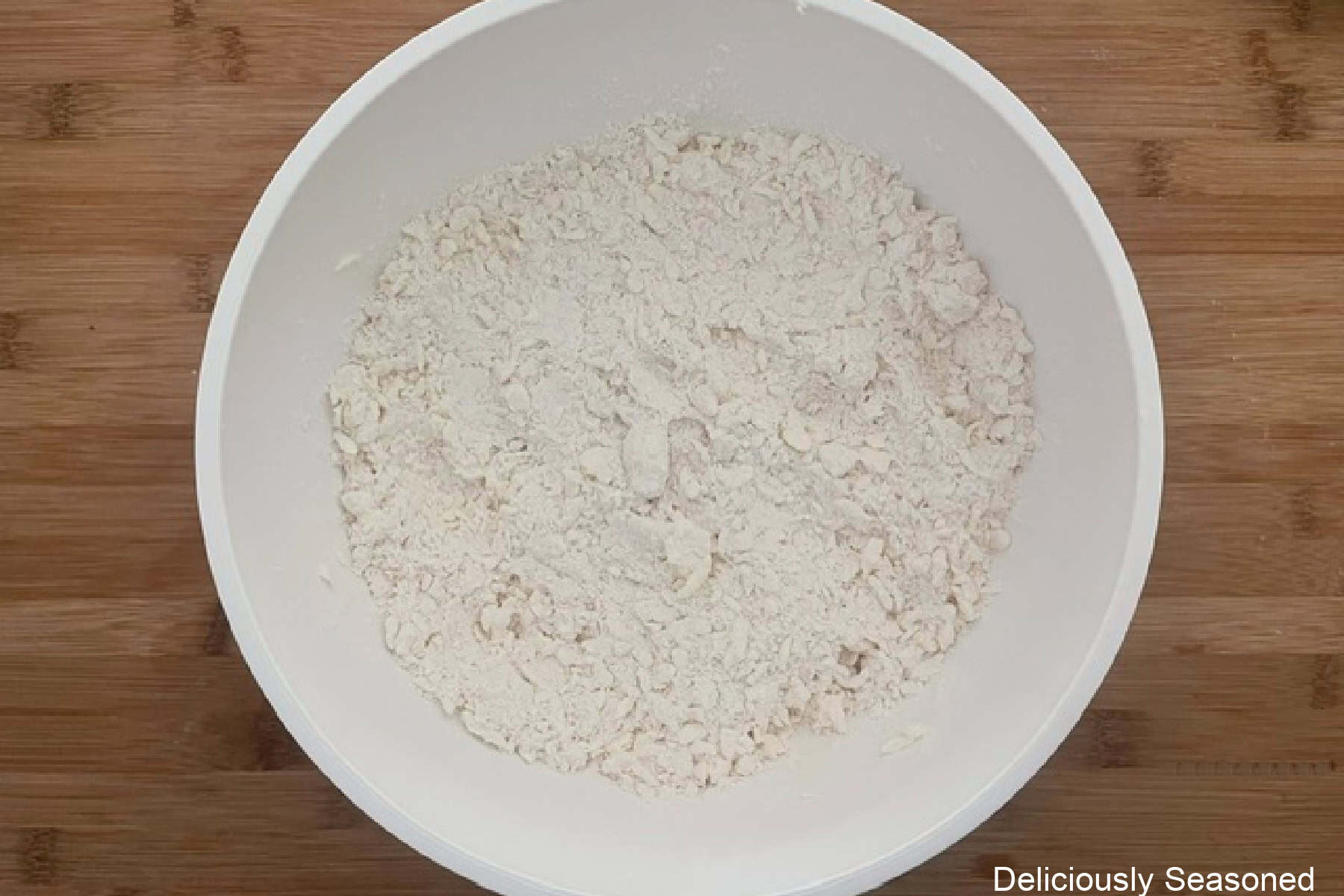A white mixing bowl filled with the flour mixture and butter mixed together, placed on a wood cutting board.