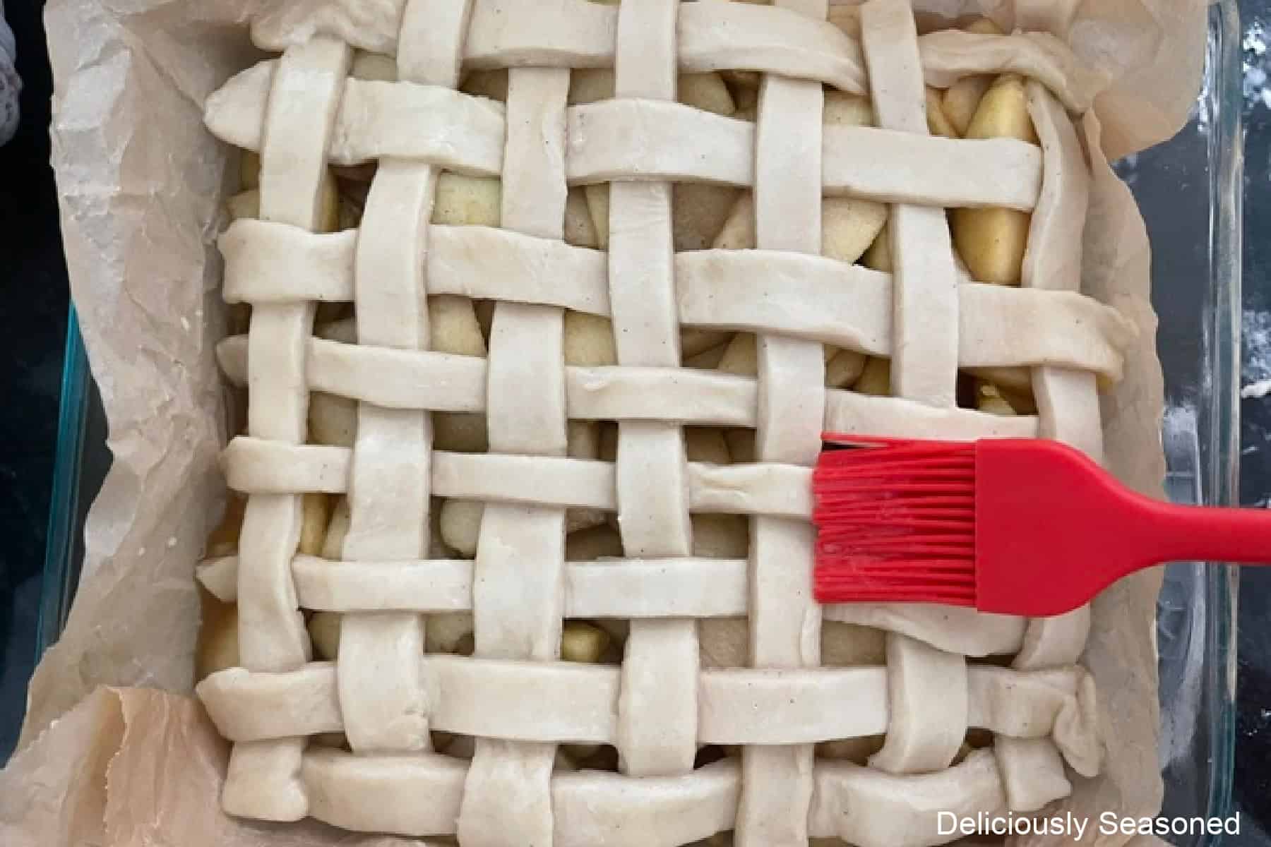 A glass baking dish with apple pie bars in it and a red basting brush brushing egg wash over a lattice topping.